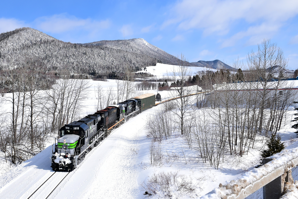 Three ex-CP RS-18's (two recently repainted into an SP Black Widow-inspired paint scheme) lead a train consisting of 72 flat cars holding 48 windmill blades (as well as 1 loaded woodchip car up front), as it approaches an overpass in the town of Carleton-sur-mer, headed for Matapedia and interchange with the CN. These windmill blades are destined for Texas and represent a new business for the Société du chemin de fer de la Gaspésie, which runs this scenic ex-CN line. While much of Eastern Canada has no snow left, there is not shortage of it out here.