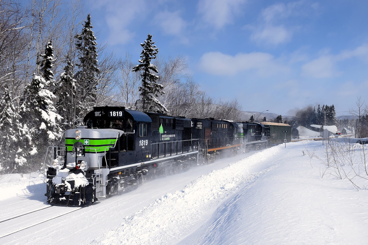 The Société du chemin de fer de la Gaspésie's windmill train is passing through the town of Escuminac, on its way to Matapedia and interchange with the CN. Power is three ex-CP RS-18's, two which have been repainted into an SP Black Widow-inspired paint scheme.
