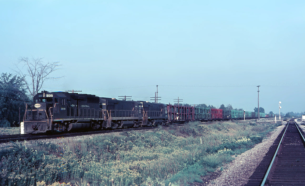 Sept 1968, an early evening finds Penn Central westbound train ML-9 ( Multi Level) passing through Canfield on the Canada Division. This train originated at Framingham MA with a day’s worth of empty racks going back to Detroit region facilities for reloading.  A GP40 and a pair of U30B’s can easily handle this train with their combined 9,000 hp but the draft created will stir up the dust and make those weeds wave back & forth for a few minutes.  That wooden property line post in the middle of the bullrushes once had CSR painted on it (Canada Southern Railway).