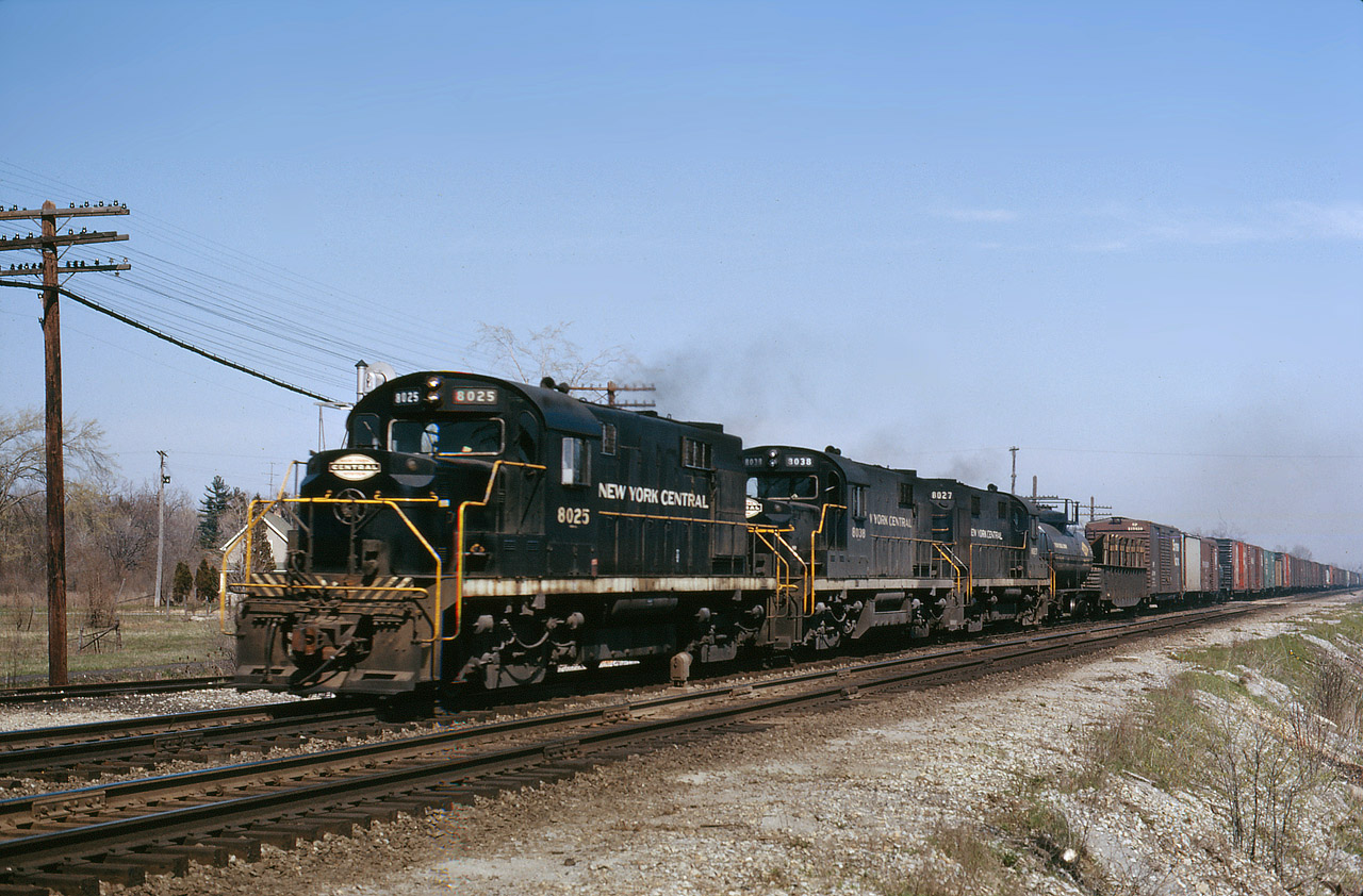 A trio of Alco RS32’s ( DL721), 8025-8038-8027, are on an NYC westbound at Canfield in May 1964. The train has a great mixture of freight including a 28-foot “shorty” tank car six cars back. The gondola appears to have a load of steel plate on a 45-degree angle, complete with bracing. The track maintenance is still being kept up, including removal of the old ties from the eastward passing track (between Doug and the road crossing). The RS32 from Alco was their counterpart to EMD’s GP20.  NYC would end up buying 25 from Alco and 15 from EMD of these 2,000 hp models. The RS32’s would get renumbered for the Penn Central merger, by changing the first digit only, from 8 to 2.