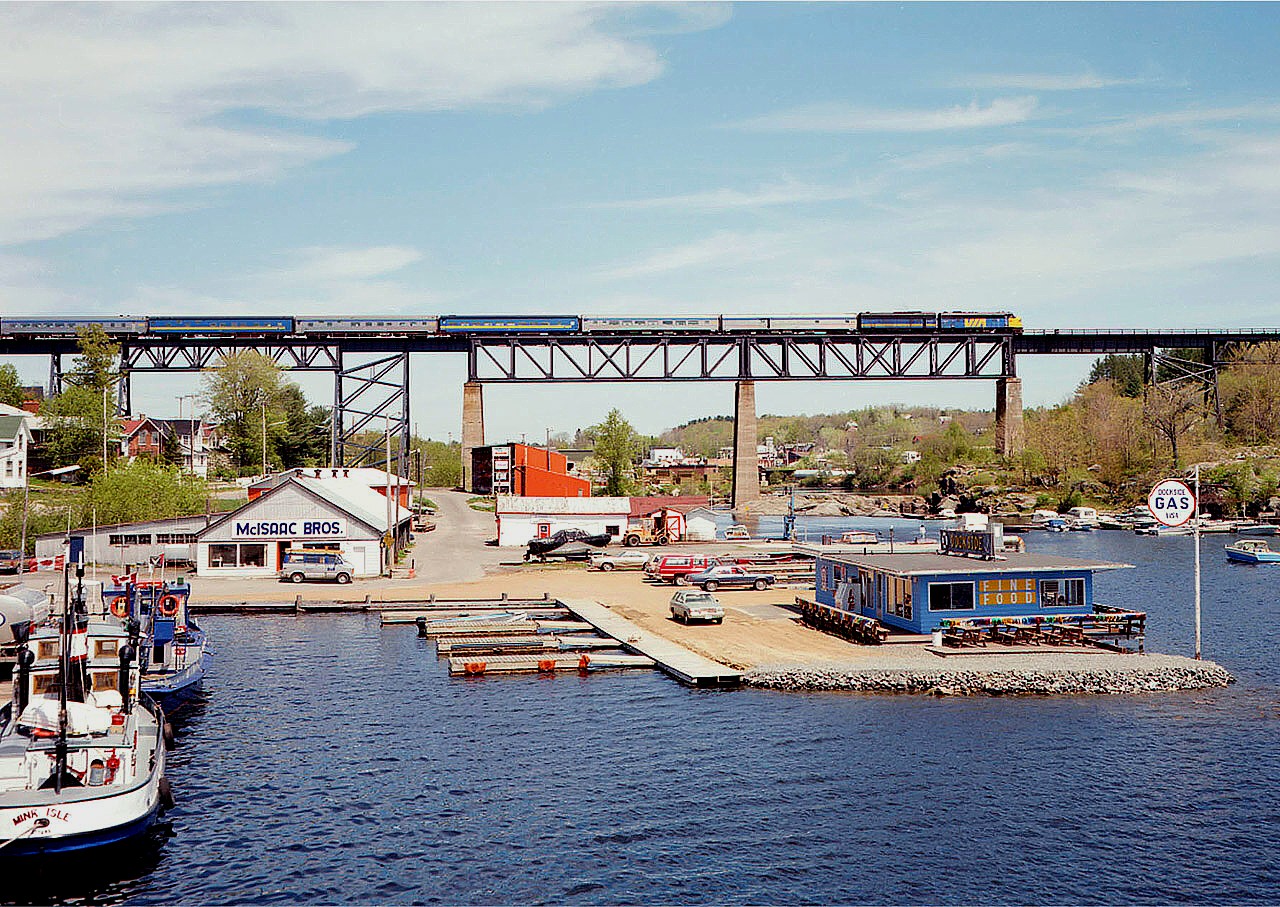 Another view of a southbound VIA over the big Trestle at Parry Sound, yes, but to this photographer it is a special image. Thus, a favourite.
It IS a beautiful sky day. I was aware of the train's approach, so I went down to the town dock and had a look around for a spot to shoot. And eyed the Island Queen, the 30,000 Islands tour ship. It was not yet into service as it is still early spring in cottage country. But there it is, and nothing but a chain to keep anyone from climbing aboard. So I climbed over, and scurried up to the top deck, and set up tripod & mount the camera. All I had to do was sit and wait in the cold on shore wind and this photo was the result. It is a nice view of the north side of the harbour as it looked back then. For the record, the power is VIA 6511 and 6636. What is unusual is the camera. At least in the world of digital this Anniversary Speed Graphic camera is a relic. I am using a Military Version,(circa 1942) coated in olive drab finish (although if one googles same, it is 'unsubstantiated'?) This is the first photo of 800 to RP I have submitted scanned directly off the 4x5 inch colour neg, and shot with Vericolor III film, 160 ISO. Set at 200 f11.  Back in 1986, this film, a box of 10 sheets cost $17.99, so already, before we even shoot we are out $1.79 per shot. Then it is $2 per sheet processing and $9 per image to print. So at $13. per photo, I am careful. But back in this era, this press camera in quality imagery in my mind could not be surpassed.