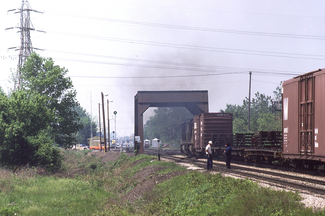 Ho hum, another day of wearing the white hat and making decisions.
I'm standing at the Sass Road crossing, recording the passage (and not-so-perfect meet) of NS train 27 with VIA number 74. Unfortunately I was not able to get the engines passing one another on this attempt, at this location. But what I did get shows a cross section of activity.
Train 27 with a pair C30-7's did not have any elevator switching to perform at Thamesville this day, so his train makeup is entirely automotive related. After passing my location, he is about to accept the CLEAR signal at the C&O diamond, as witnessed by the double greens on the dwarf signal, just ahead of the lead unit.
The high green signal is the Clear train order board at Chatham East. And except for the two fellows nearest my lens, all other personnel are standing off to the south of the eastbound main as VIA 74 looms in the distance.
Taken with a Hanimex 300mm mounted on a Canon FTb.