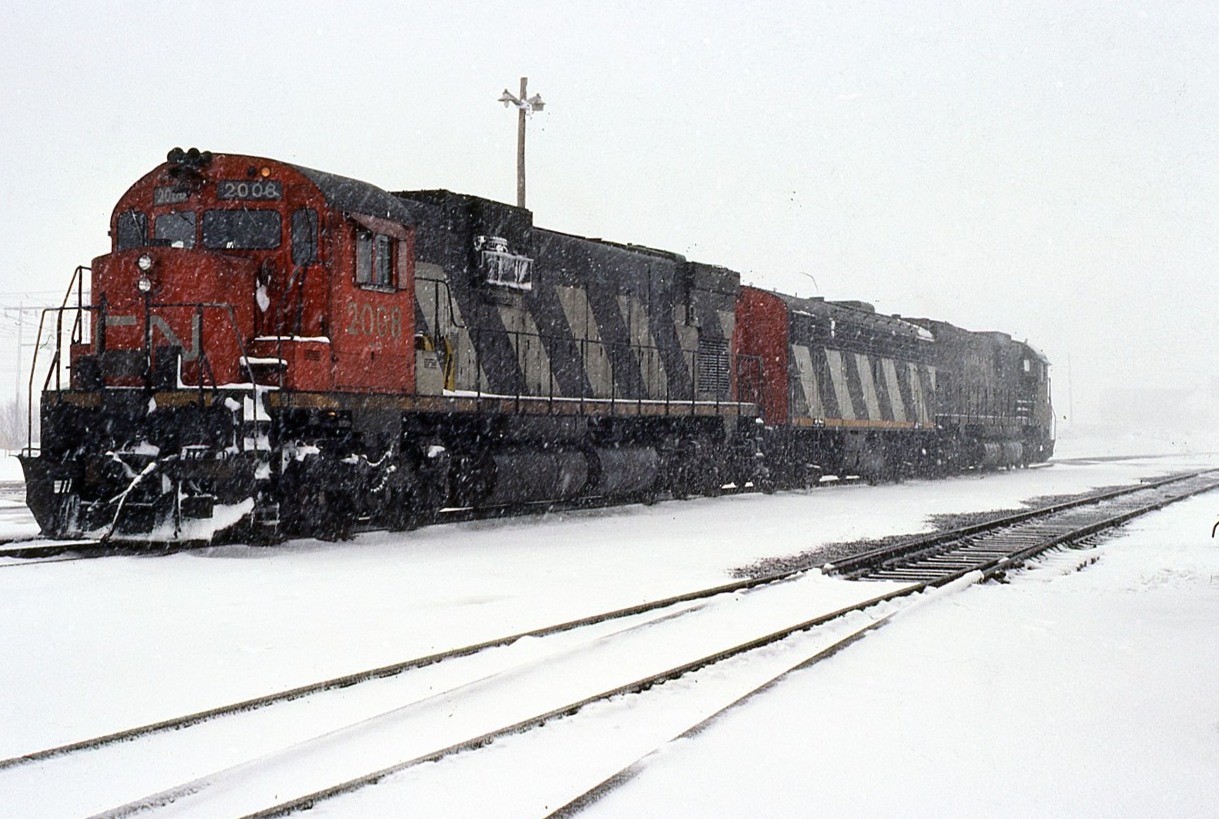 A Ron Hagemeister photo.
During a heavy spring squall, 3 freight engines wait for the call to duty. A pair of C630's are bracketing F7B 9191 near the east end of Sarnia Yard.