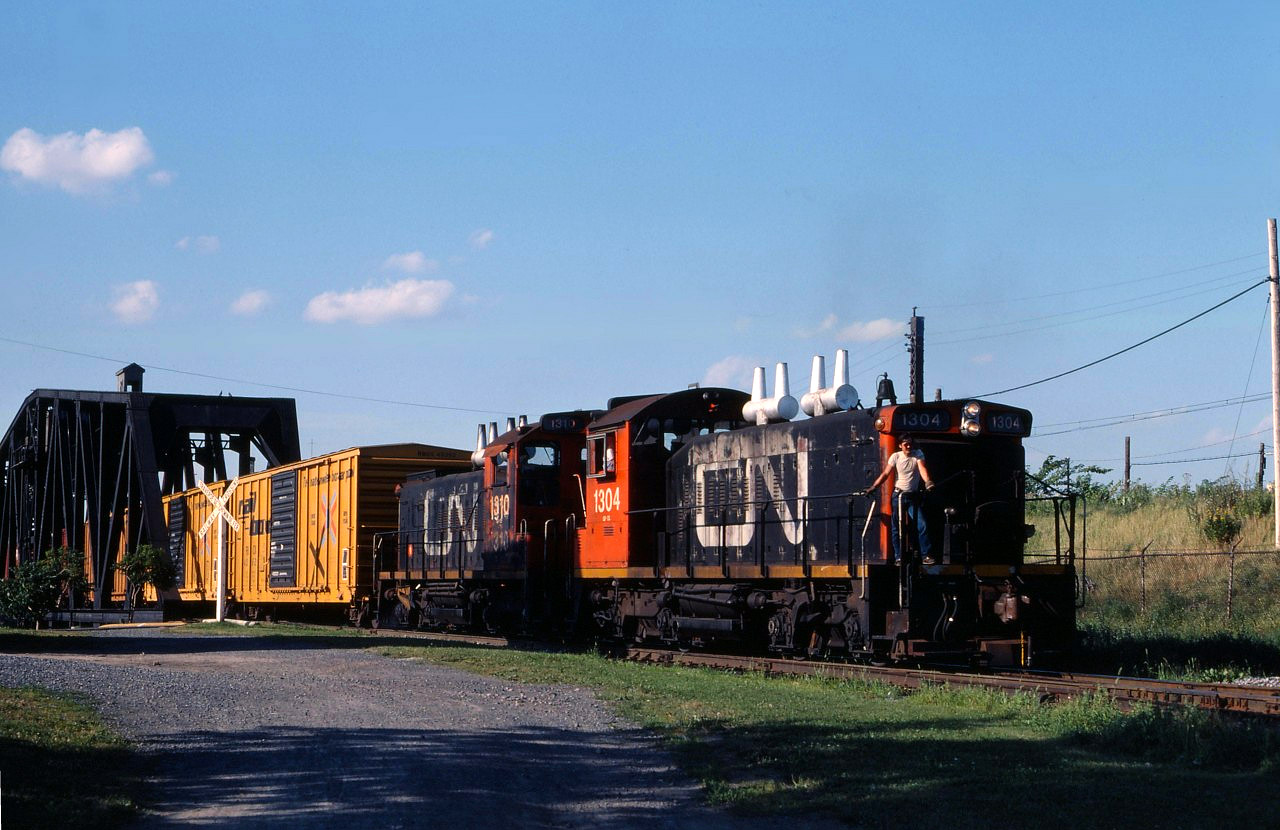 A crew member watches for pedestrians and cyclists, and prepares to flag crossings up ahead, as a couple of "pups" (SW1200RS units) cross the Lachine Canal near Wellington Tower in 1985. The train is returning with a transfer from the National Harbours Board.