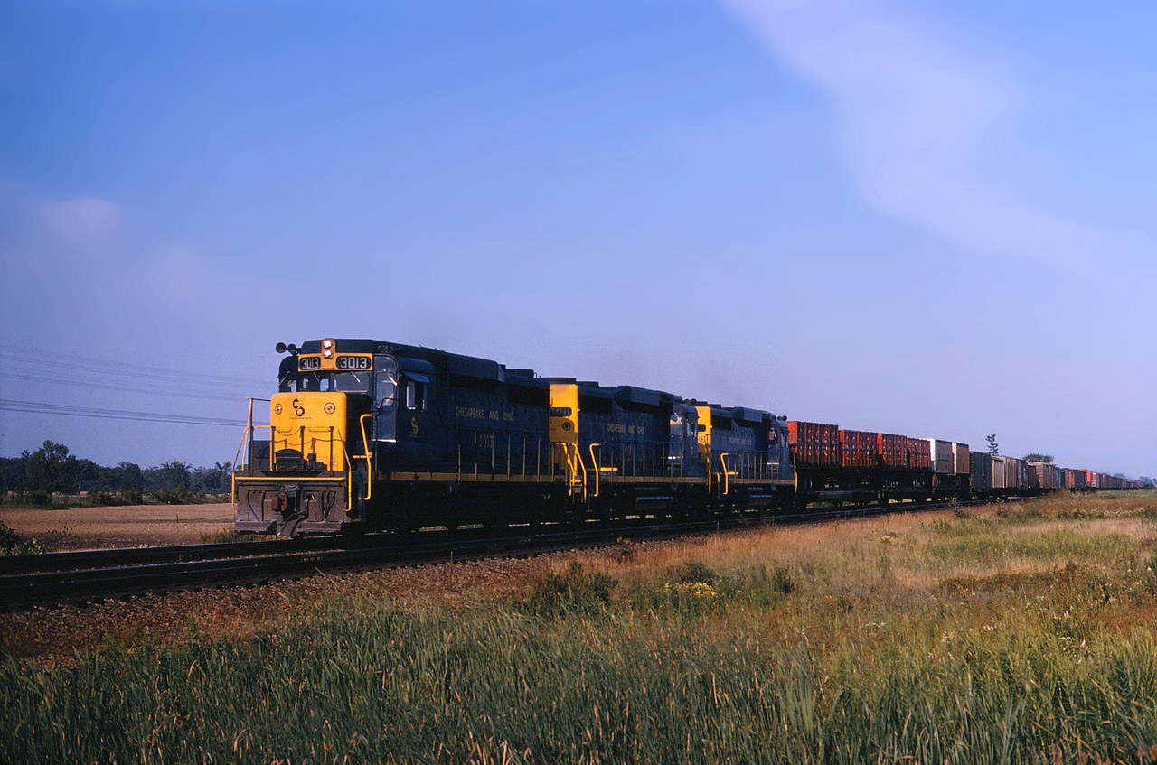 Another trio of fairly new GP30’s has a westbound C&O freight moving towards St. Thomas where some of the cars will go further west to Detroit, and others will go north to Sarnia, Port Huron and beyond.
There are some interesting cars in the train today, starting with specialty containers on flatbed trailers right next to the power. I believe this photo was taken at the highway 3 crossing, facing east. The Cayuga Sub would be just out of the picture and you can see at least one of the wooden property line posts placed in the swamp  by Canada Southern Railway. Plus the train is coming over a slight rise which is how the right of way looked at this location. Highway 3 near the junction of 56 is in the background.