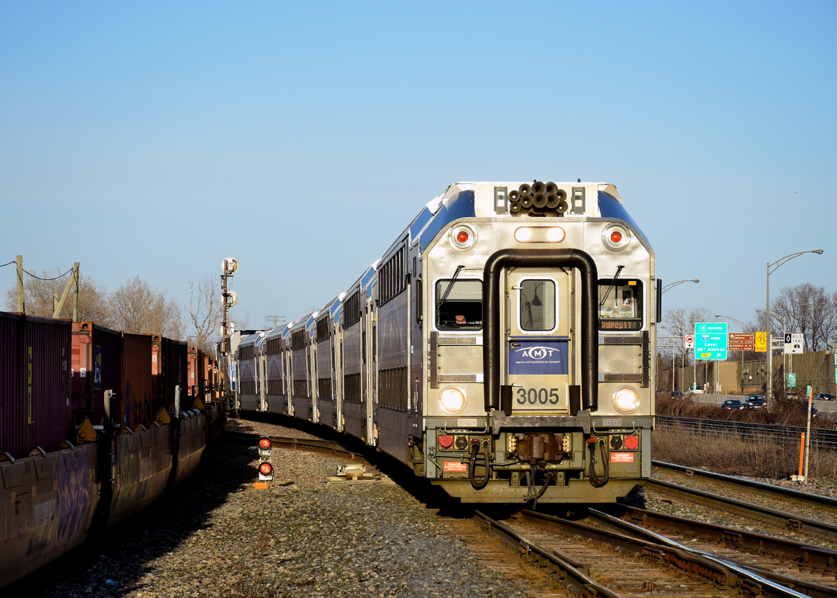 AMT 29 with cab car AMT 3005 is approaching its stop at Lachine Station at the end of a sunny day. Intermodal train CP 143 at left slowly passes at left, once AMT 29 leaves the station CP 143 will follow it westwards along the Vaudreuil Sub.