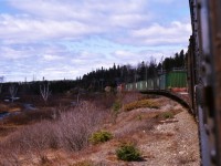 BADGER BOUND. The sun breaks through just in time on the bright green Terra Transport inter-modal containers as Mixed Extra 945 West heads towards the town of Badger on April 20, 1987. Having just left Grand Falls on his first Newfoundland train ride and accompanied by his girlfriend, the photographer opened the window on Coach 757 and carefully held his Pentax ME-Super to capture this scene. For the remainder of the journey, all photos were shot leaning out the vestibule as the trainmen had kindly given permission that it was OK to do so. At this point, the train ran parallel to both the TCH and the Exploits River but at Badger the highway would turn north and it would not be seen again from the train until Deer Lake, some 80 miles west. It would also be the last views of the Newfoundland spring until then as the climb into the Gaff Topsails would bring back the still remaining winter vistas.