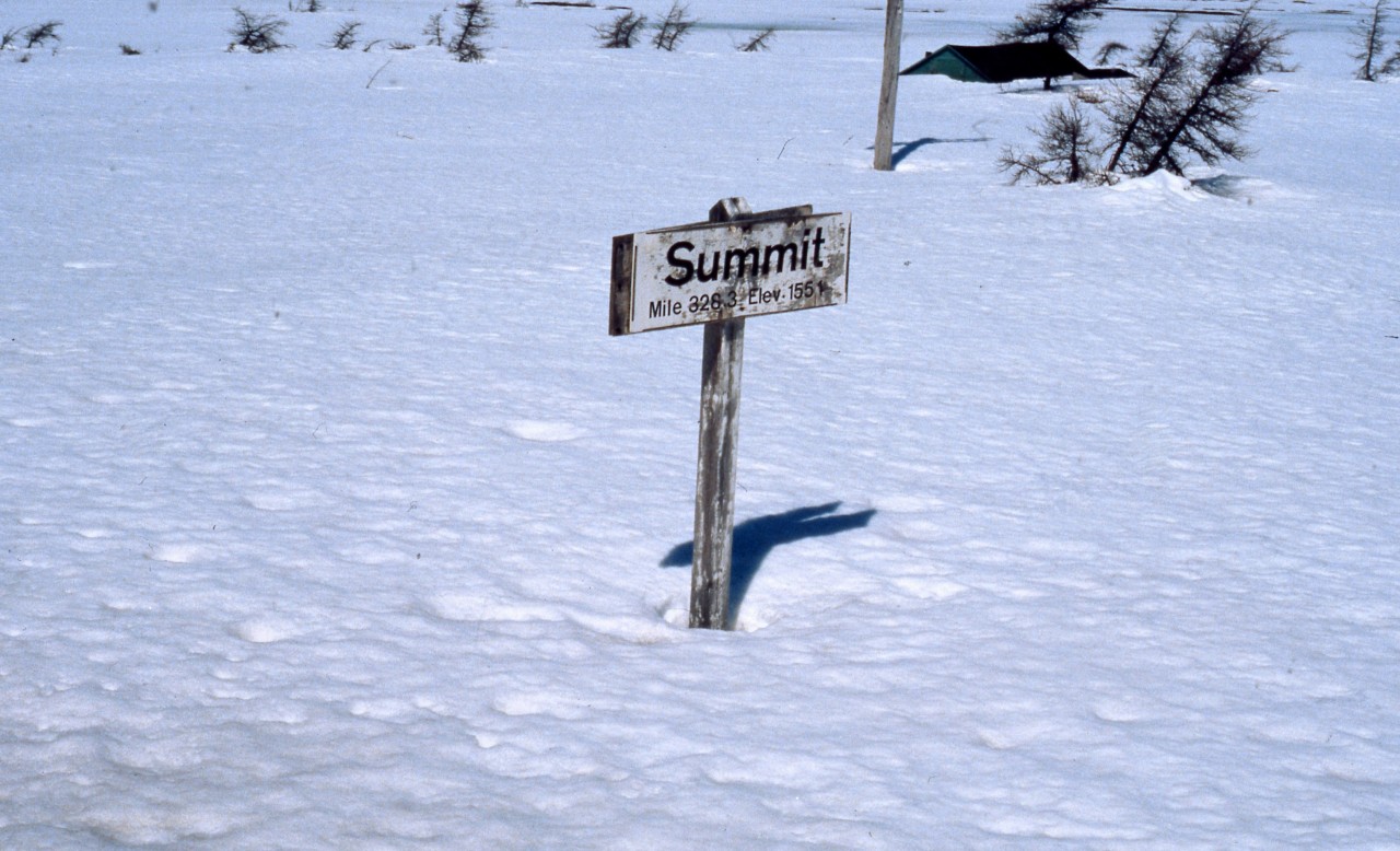 SIGNAGE AT SUMMIT. As seen from Coach 757 on Terra Transport's Mixed Extra 945 West on April 20, 1987, the official sign for 'Summit' is barely visible above what remains of a heavy winter's snowfall. It was at this point at Mileage 328.3 that the former Newfoundland Railway reached it's pinnacle at 1554 feet above sea level and no doubt that a few months earlier, the sign would have been completely covered under snow. While the rugged Juniper trees point east as part of their distinctive growth pattern, the photographer and his girlfriend continue to head west on this, their first Newfoundland rail journey. This sign has long since disappeared, along with the other official markings of the 547 mile narrow gauge railway, but has been replaced by a man-made one that not only displays the mileage and elevation in raised lettering but also wooden representations of the Canadian and Newfoundland & Labrador Flags.