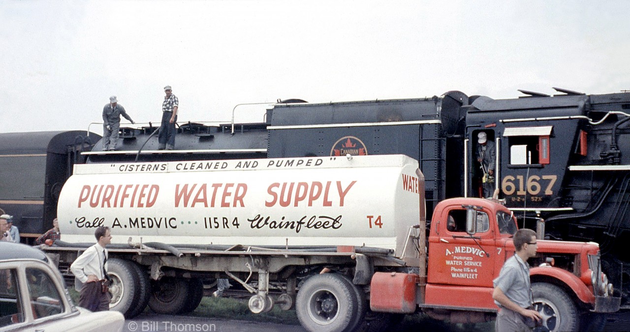 Fantrip attendees and railfans bearing cameras are stretching their legs during a stopover at Port Colborne on the July 10th 1960 UCRS fantrip. The crew of CNR Northern 6167 attend to her needs as she takes on water from one of A. Medvic of Wainfleet's tanker trucks.