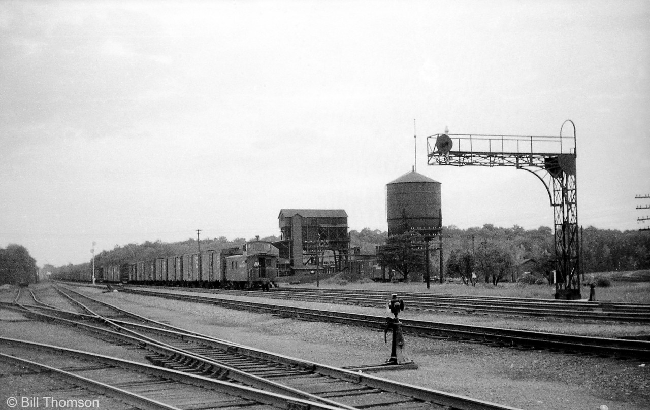 A general overview of Canadian Pacific's Guelph Junction is shown, looking west along the Galt Sub in August 1959. The tail-end of a westbound freight is shown, along with the westbound signals, water tower, and coal tower. The shop building is hidden in the background, and the station is out of view just to the right of the photo. The CP Goderich Sub to Hamilton (presently part of the Hamilton Sub) branches out at the bottom left, with the north portion of the Goderich Sub continuing to Guelph (the Guelph Junction Railway) and on to Goderich partially visible in the background on the right.