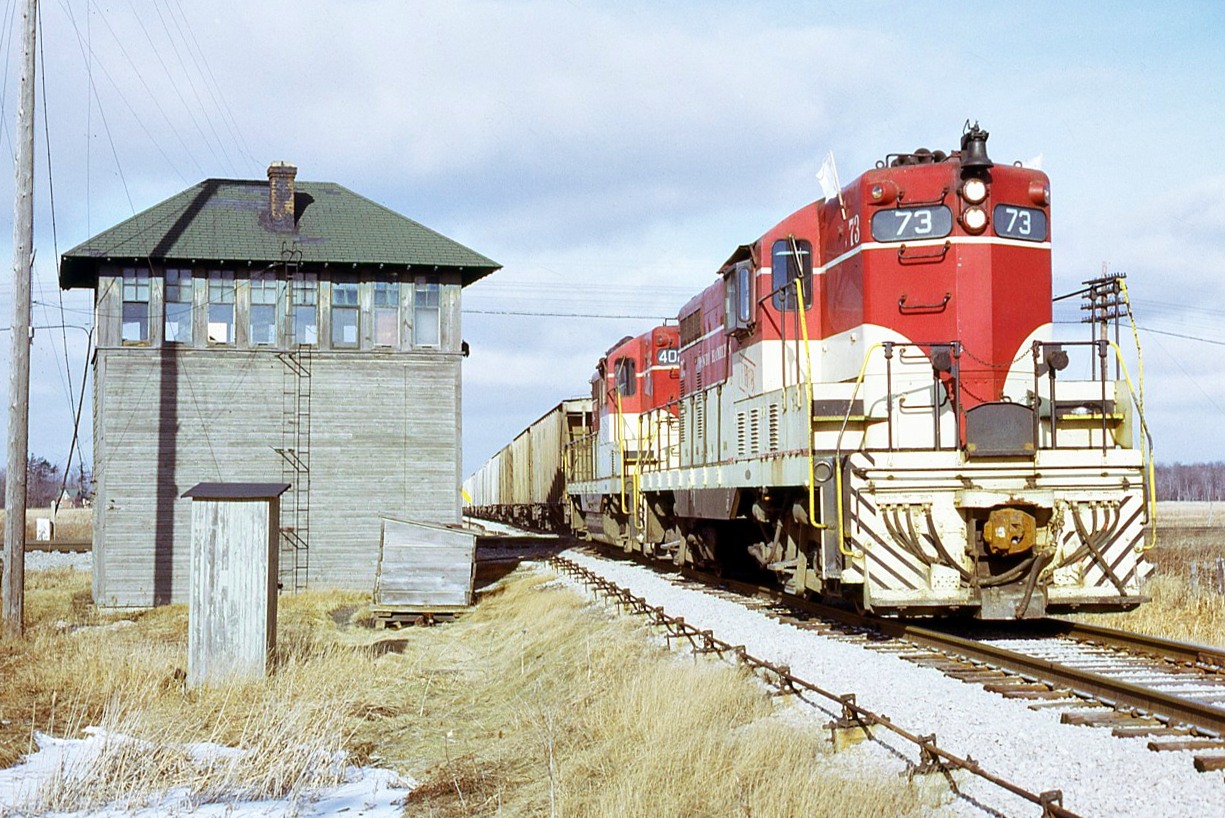 TH&B 73 and 402 are southbound on the Dunnville Subdivision with over 45 cars of phosphate rock for ERCO at Port Maitland. They are passing by Penn Central's 'E&O' interlocking tower as they clunk-clunk across the Canada Division mainline. PC employee, Operator/Leverman WA Webster has locked the armstrong levers into proper position to allow for this movement. The 3 wooden structures ( tower, outhouse and coal bin) are well weathered at this stage.
