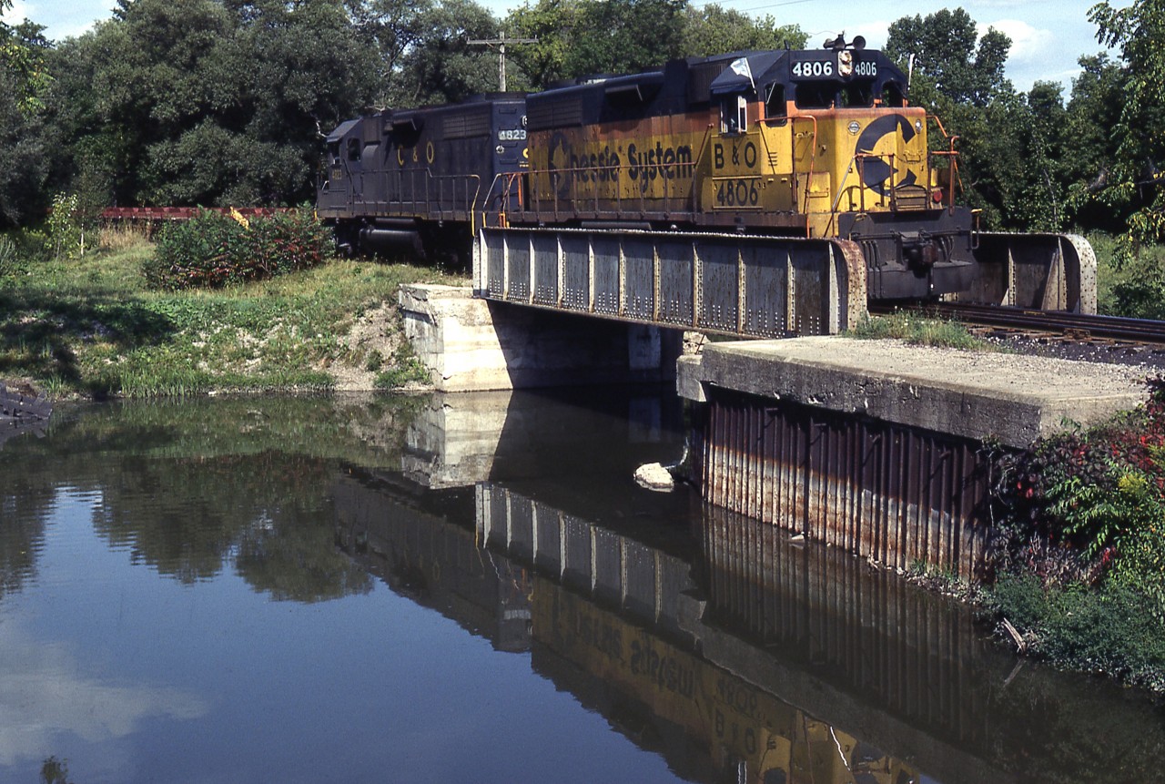 With leased GP38's reflecting in the water, Extra B&O 4806 West has descended the grade off the Cainsville Fill and is about to cross Locks Road in the southeast end of Brantford. This is where water collected in Mohawk Lake drains via a small damn into the Grand River. On this day the empty slab cars are right on the head end. You can visit this location via the Hamilton-Brantford Rail Trail,  which utilizes the TH&B right-of-way.