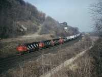 Westbound CN #425 with a long string of TOFC up front, something we rarely see any more, climbs the grade at mile 4.4 Dundas. The remnants of old Canada Crushed Stone operation can be seen on the hillside. (This business shut down around 1974) A siding on the south side is still in, MoW used this track frequently. In the lower level, the lead to the old Steetley dolomite operations (as well as one time a TH&B connection) was taken up in October 1985. That is Sydenham Rd bridge in the background. Power shown is CN 9507, 9411, 9501 and 2108. Photo shot with Speed Graphic 4x5 at 1000 f5.6 and 160 ISO Vericolor III film. This nice vantage point was overgrown by relentless foliage many years ago, unfortunately.