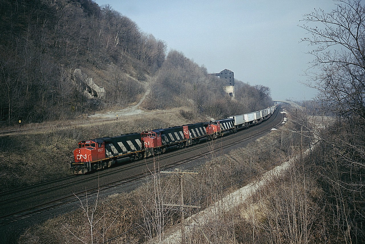 Westbound CN #425 with a long string of TOFC up front, something we rarely see any more, climbs the grade at mile 4.4 Dundas. The remnants of old Canada Crushed Stone operation can be seen on the hillside. (This business shut down around 1974) A siding on the south side is still in, MoW used this track frequently. In the lower level, the lead to the old Steetley dolomite operations (as well as one time a TH&B connection) was taken up in October 1985. That is Sydenham Rd bridge in the background. Power shown is CN 9507, 9411, 9501 and 2108. Photo shot with Speed Graphic 4x5 at 1000 f5.6 and 160 ISO Vericolor III film. This nice vantage point was overgrown by relentless foliage many years ago, unfortunately.