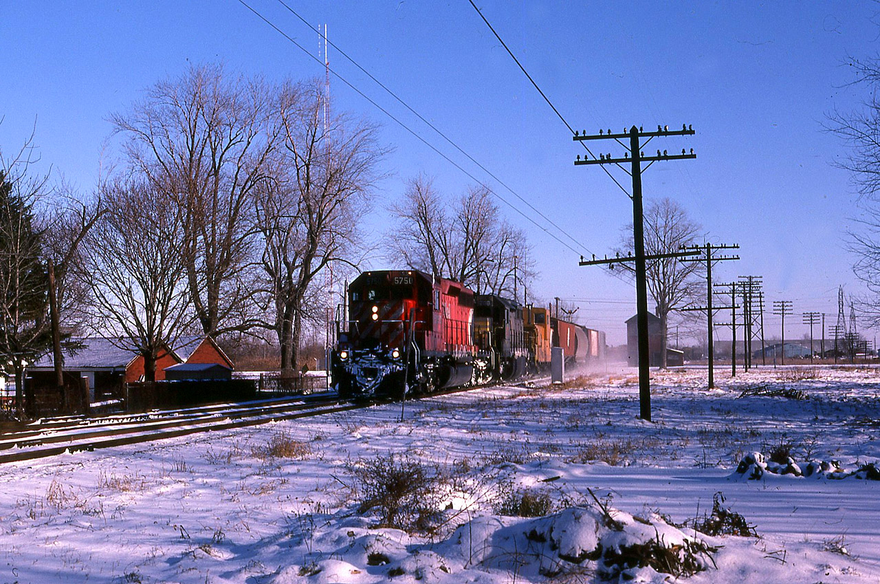 A cool shot on a bitterly cold day finds CP westbound time freight 917 with 5750- QNSL 208 (being purchased by CP) making a small setoff at Chatham.
There was an expression used by C&O workers, that on cold winter days when the snow is dry, it " runs " as the wind pushes it from one place to another. This was in reference to otherwise flat terrain where the snow would 'run' and accumulate to the lee of a hedgerow or a stand of trees. Such is the case on this bright morning, as 917 appears to be moving at track speed. In fact, the head-end man has made the cut on the Chatham cars and they are pulling ahead to clear the plant, in order to get the rail & signal to shove the cars into the yard. The caboose was probably being changed out for the Chatham-based roadswitcher.