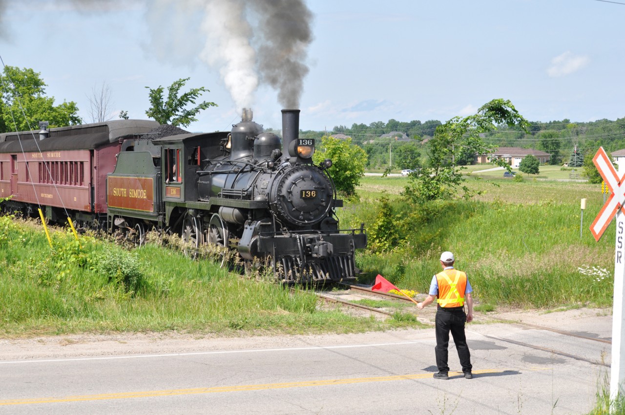 A crossing guard stands watch as the engineer of South Simcoe Railway 136 sounds the whistle. She is seen here crossing the 5th line, and heading south on the old Beeton Sub to Tottenham station to finish the day.