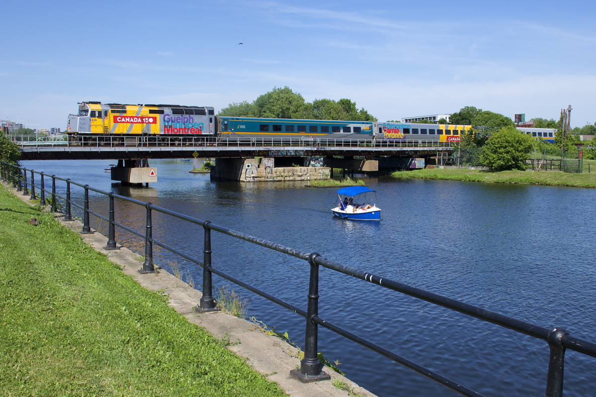 VIA 67 has LRC cars in three paint schemes and wrapped VIA 6436 leading as it crosses the Lachine Canal, with a pleasure boat passing at right.