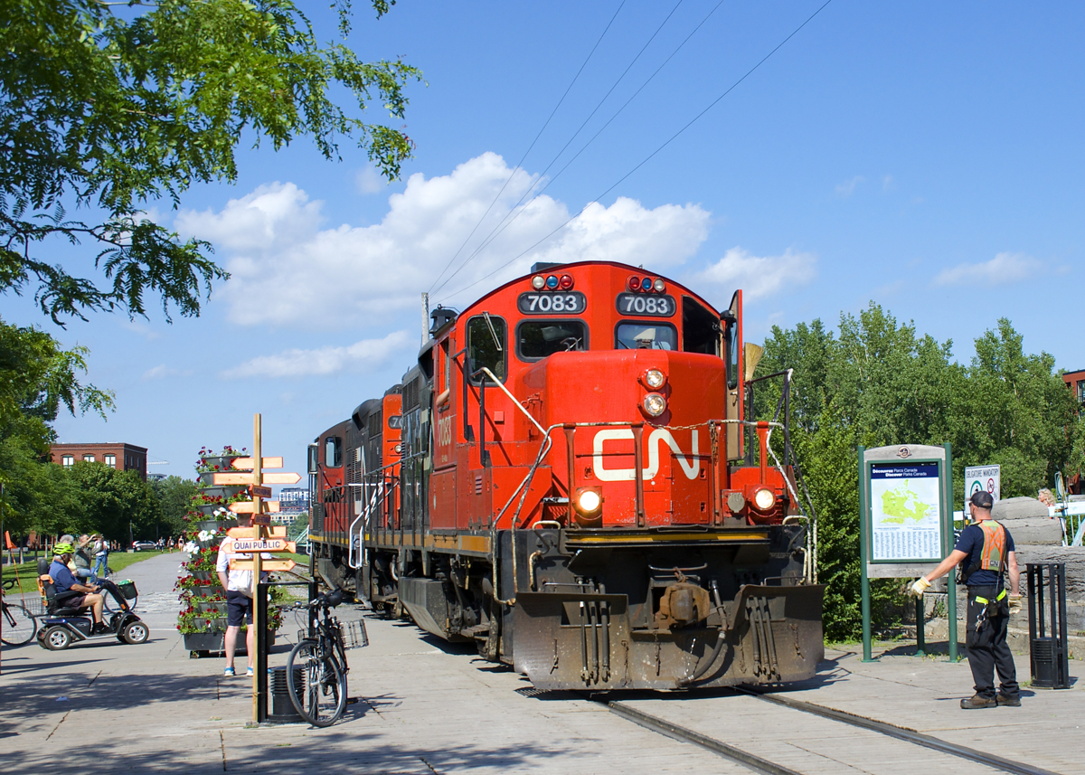 A crewmember is flagging a pedestrian crossing beside the Atwater Market as a pair of GP9's (CN 7083 & CN 7054) head light on the East Side Canal Bank Spur after dropping off eight cars at the Robin Hood Flour Mill (the last client on this line).
