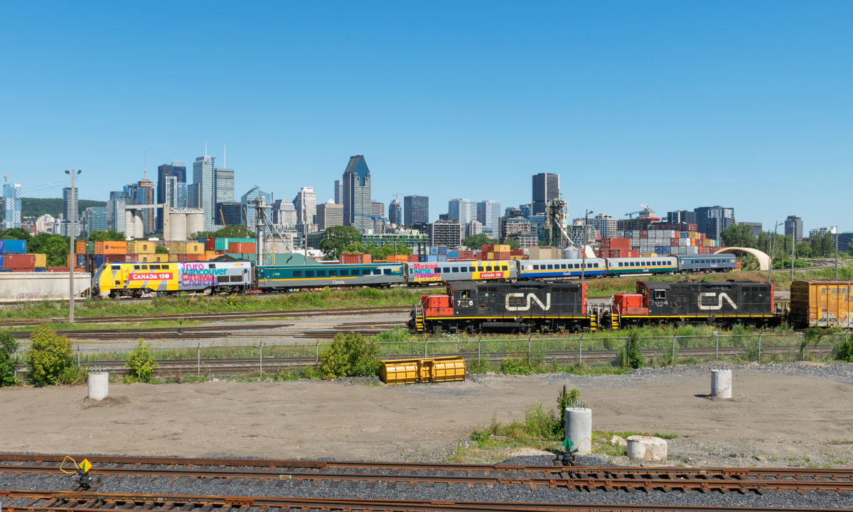 VIA 65 with wrapped 913 and a wrapped car (that lists Montreal on its side, along with Sarnia, Winnipeg & Alexandria) passes the Pointe St-Charles Yard, where a trio of GP9's (CN 7226, CN 7054 & CN 7083) are switching the yard.