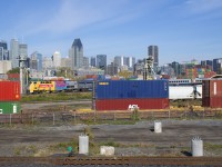 The lead unit of VIA 65 (VIA 907) is seen between containers heading east on CN 120, as both pass through the Pointe St-Charles neighbourhood of Montreal, with downtown Montreal in the background.