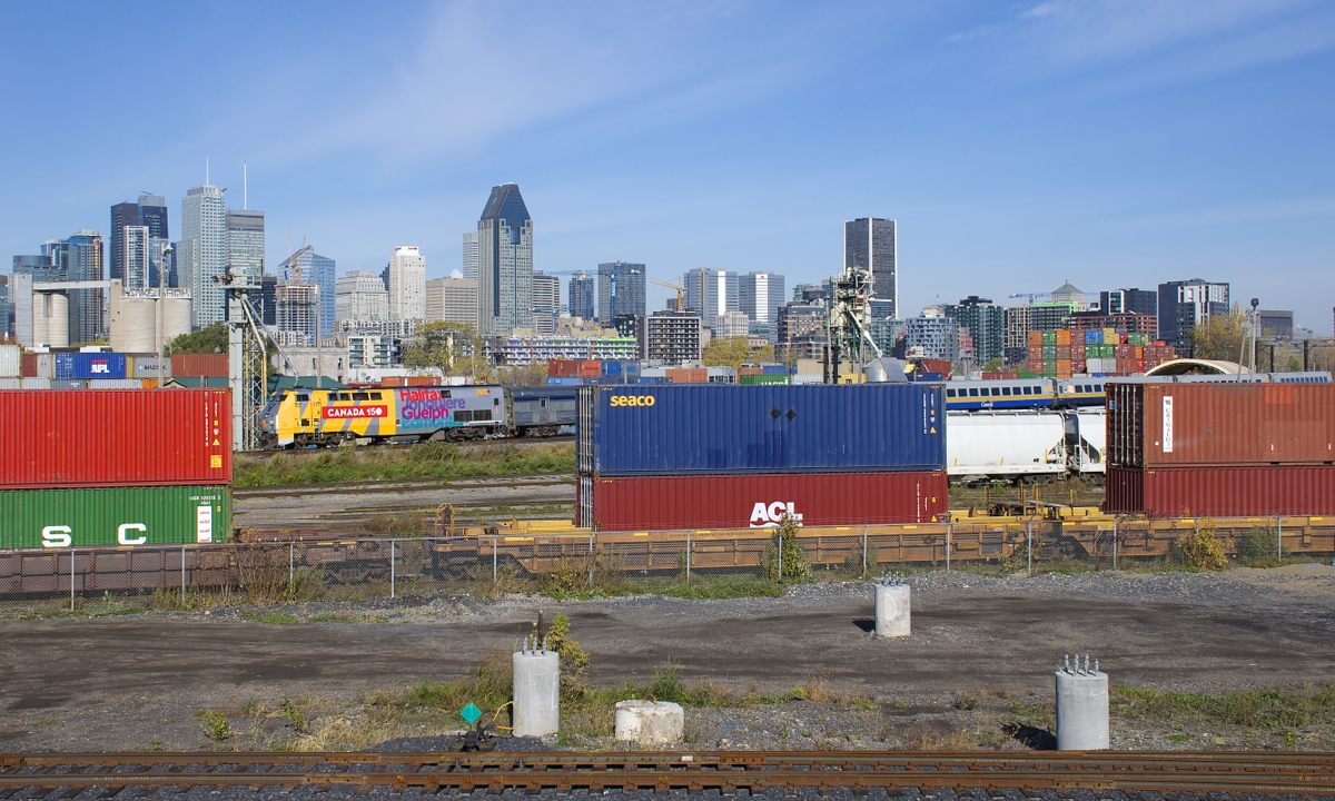 The lead unit of VIA 65 (VIA 907) is seen between containers heading east on CN 120, as both pass through the Pointe St-Charles neighbourhood of Montreal, with downtown Montreal in the background.