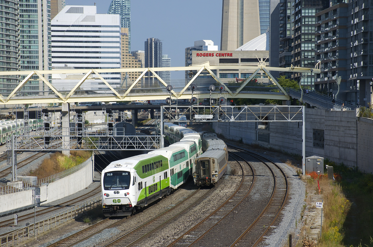 A GO Transit and a VIA Rail train are both heading east towards Union Station as they pass under an older signal bridge with classic searchlight signals. At left is a new signal bridge, not yet in use.