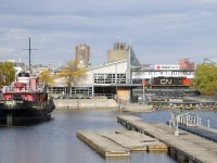 While waiting for security to arrive and flag crossings in the Port of Montreal, CN 7228 is stopped on the bridge over the Lachine Canal, out of sight at its left is slug CN 276 and a single tank car. The tank car is destined for interchange the Port of Montreal, with the power shoving the car in. The tops of a number of Montreal structures are barely visible in the distance, from left to right are Bonsecours Market, the Olympic Stadium, the Radio-Canada building, the Molson brewery and the Jacques-Cartier bridge. In the foreground is the preserved tug <i>Daniel McAllister</i>.
