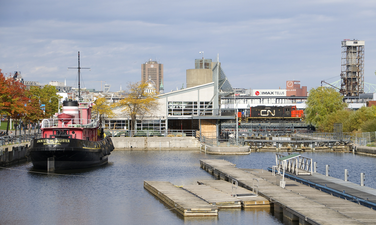 While waiting for security to arrive and flag crossings in the Port of Montreal, CN 7228 is stopped on the bridge over the Lachine Canal, out of sight at its left is slug CN 276 and a single tank car. The tank car is destined for interchange the Port of Montreal, with the power shoving the car in. The tops of a number of Montreal structures are barely visible in the distance, from left to right are Bonsecours Market, the Olympic Stadium, the Radio-Canada building, the Molson brewery and the Jacques-Cartier bridge. In the foreground is the preserved tug Daniel McAllister.