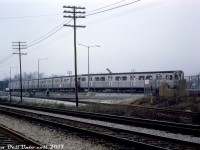 A 4-car set of Montreal Locomotive Works (MLW) built "M1" subway cars sits at the back of the TTC's new Greenwood Yard in storage, as viewed from CN's Kingston Sub mainline to the south. Greenwood Yard was built in the 1960's in Toronto's east end to store, maintain and service trains on the under-construction Bloor-Danforth subway line. The opening of the B-D line was still 3 months away (February 1966), and Greenwood wasn't fully operating yet, but since May it had been taking deliveries by rail of new H1 subway cars that would provide service on the line.
<br><br>
The M1's (numbered 5300-5335) were the first new subway cars built in Canada, and featured a 75-foot light-weight aluminum bodied design that's basically still the standard for new TTC subway cars today (compared to the original steel 57' "Red Rocket" Gloucester cars made in the UK). They were built a few years earlier to serve on the then-new University subway line. MLW only got one order from the TTC in the early 60's, all subsequent orders of the same design went to Hawker Siddeley Canada (H1, H2, etc). Due to their historical significance, the first two cars of this order (M1's 5300 & 5301) were donated to the Halton County Radial Railway museum for preservation when the series were being retired in the late 90's.
<br><br>
The standard-gauge railway siding (or spur) into the yard had two legs that formed a C-shape, visible in this photo and in <a href=http://jpeg2000.eloquent-systems.com/toronto.html?image=ser12/s0012_fl1966_it0047.jp2><b>aerial imagery</b></a>. The western leg went to the unloading ramp that was used to unload new subway cars that were delivered by rail flatcar. Another siding looped up by one of buildings on the south-east side (perhaps for delivering materials). The first deliveries by rail here were the H1 cars in 1965, and the final ones were apparently the T1's in the late 90's/early 2000's. Some of the siding is still intact but out of use (with the switch to the mainline removed), and all current subway car deliveries are trucked down from Bombardier in Thunder Bay.
<br><br>
<i>Robert D. McMann photo (slide unnamed, but likely), Kodachrome from the Dan Dell'Unto collection.</i>