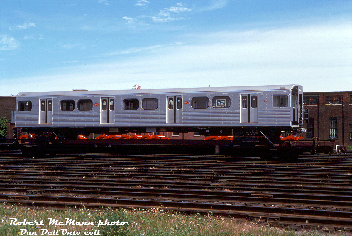 Built new for the Toronto Transit Commission in the mid-70's by Hawker Siddeley Canada in Thunder Bay, brand new TTC "H5" subway car 5741 sits on a NIFX 1000-series flatcar at Canadian Pacific's Parkdale Yard, awaiting interchange to CN and final delivery in east end Toronto to the TTC's Greenwood Yard via spur track. Note the red farm equipment on flatcars behind, from the nearby Massey Ferguson factories in the industrial Liberty Village part of town (since gentrified greatly with condos and lofts).The 138-car group of H5's was just the latest iteration of the basic RT-75 75' long aluminum bodied subway car design introduced by MLW in the early 60's as the M1, and followed by HSC's H1, H2, H3 & H4. Changes included smooth end top cap pieces by the top lights (instead of profiled like on the H1-H4), "Chopper Control" (for stepless acceleration and regenerative braking) and were the first cars to feature air conditioning on the system. The H5's were also initially delivered with "blackface" black ends, but were eventually all repainted silver. Known by passengers for their yellow doors, whining noise when accelerating or braking (from the chopper control), and musty air conditioning condenser smell, the H5's were phased out by the new Bombardier Toronto Rocket trains starting in the early 2010's, and the final train of H5's made its last run on the Yonge-University-Spadina line in June of 2013. Most were supposed to be refurbished for use on a Lagos light rail project in Nigeria, and many were shipped to New York for refurbishing, but the plan fell through (new rolling stock was later purchased) and the H5's were cut up for scrap. Apparently some Canadian enthusiasts were able to contact the scrapper in the US, and obtained a U-haul load stash of H5 goodies, but that's another story...For the rail enthusiast, the eclectic mix of rolling stock used to ship subway equipment varied over the years, from standard CP flatcars used to deliver the original 57' long "Red Rocket" Gloucester cars in the 50's, specially built CP 6-axle flatcars made from cut down heavyweight passenger cars, to these NIFX 1000-series 4-axle flatcars equipped with pedestal supports (originally lettered "North American Car Corporation", some painted out to leave "Can Car", presumably leased to HSC/Can-Car in Thunder Bay), to yellow 89' TTX flats used to ship the H6's. The last subway deliveries by rail were the T1's to Greenwood in the 90's, and all subway cars in and out of the system are presently moved by truck and special low-loader flatbed trailer.Robert D. McMann photo, Kodachrome from the Dan Dell'Unto collection.