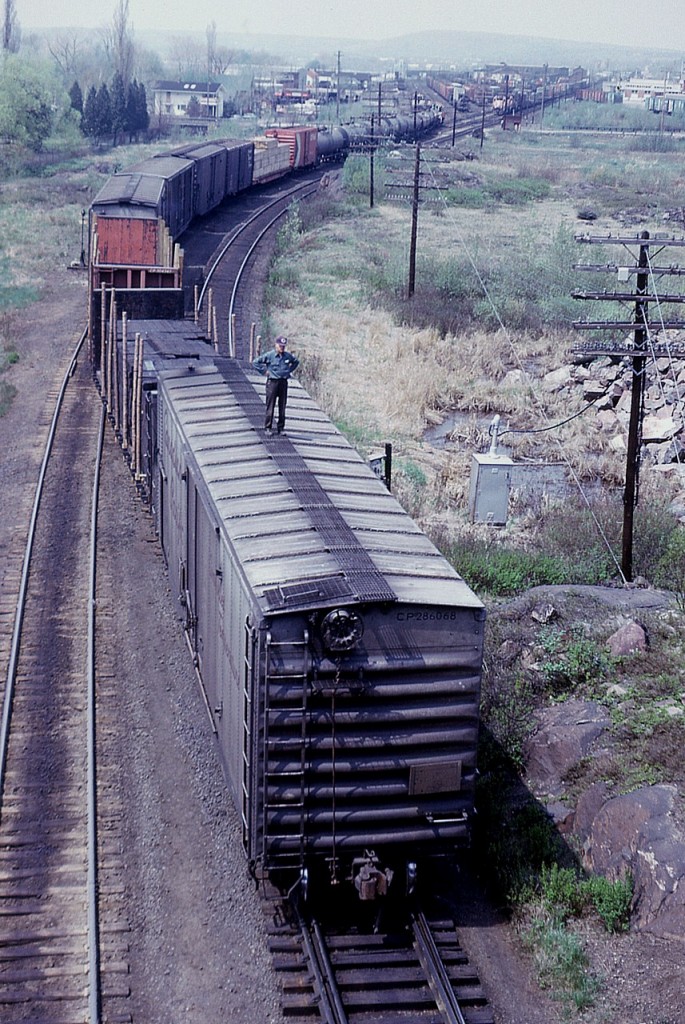 "Be Safe Out There"!!   Sure appears safety rules are being compromised in this scene, but then, not ever being an employee of the railroads I do not know what rules were in place back in 1975 and perhaps this was a normal procedure; riding the car. I'd like to know when changes took place. Certainly looks dangerous to me, and the 'coupling up' had better be gentle!! It is a misty weekend in North Bay, and a hot one, as I remember. That is the CP yard in the background, the main city downtown in the distance on the right.