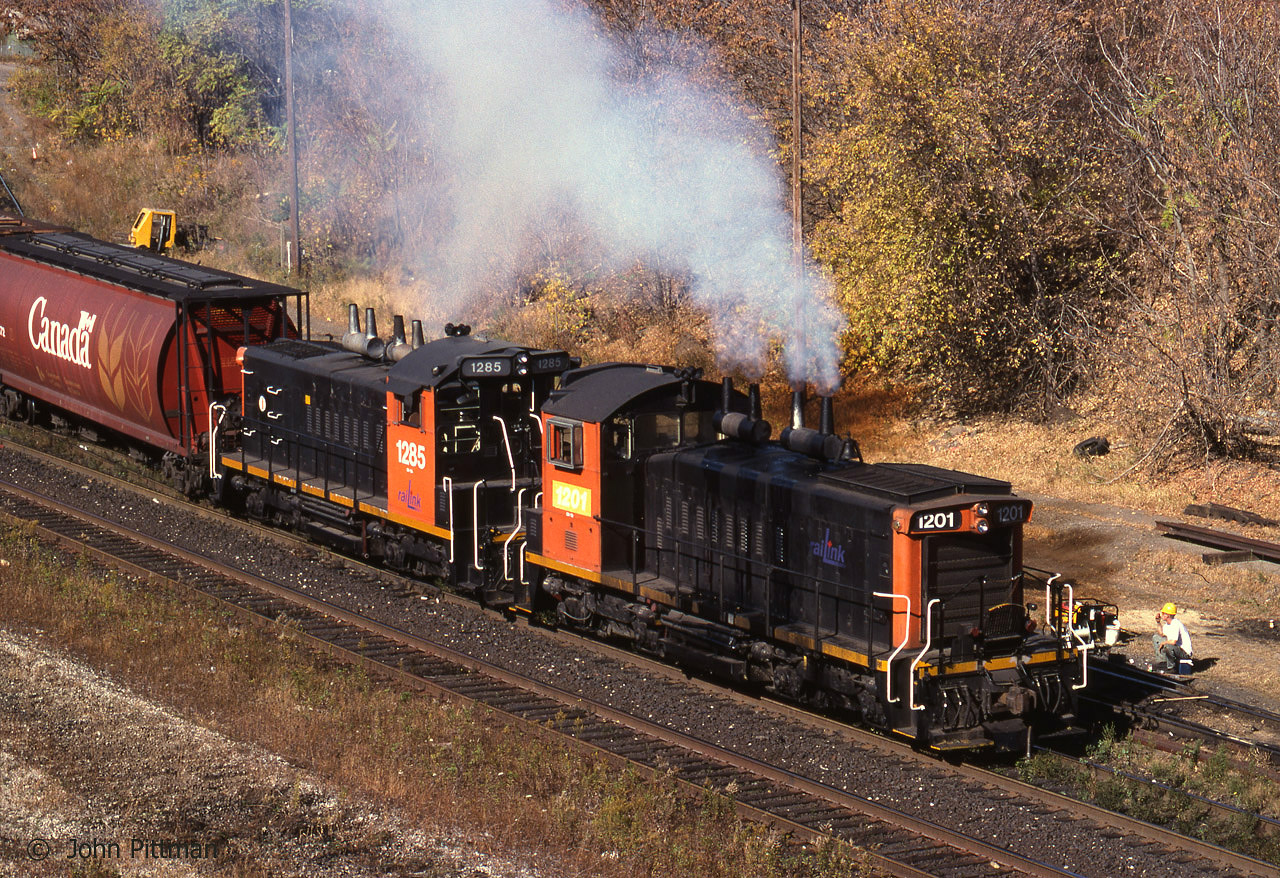 First autumn of RaiLink Southern Ontario Railway operation of CN's Stuart Street Yard on the west side of  Hamilton ON.  Two elderly SW1200RS work the yard, the first RaiLink units I ever met (seen in Brantford Yard before their first day in Hamilton).  The earlier style cab numberboard & headlight fairing of ex-CN 1285 differ from those of 1201 which had its original CN 1335 number replaced, to reflect its horsepower most likely.  Note the fuel stains from 1201's stacks, and the "speeder" beyond the wheat hopper car.  It appears that the guy wearing a yellow hardhat is taking a break from a maintenance of way job.
