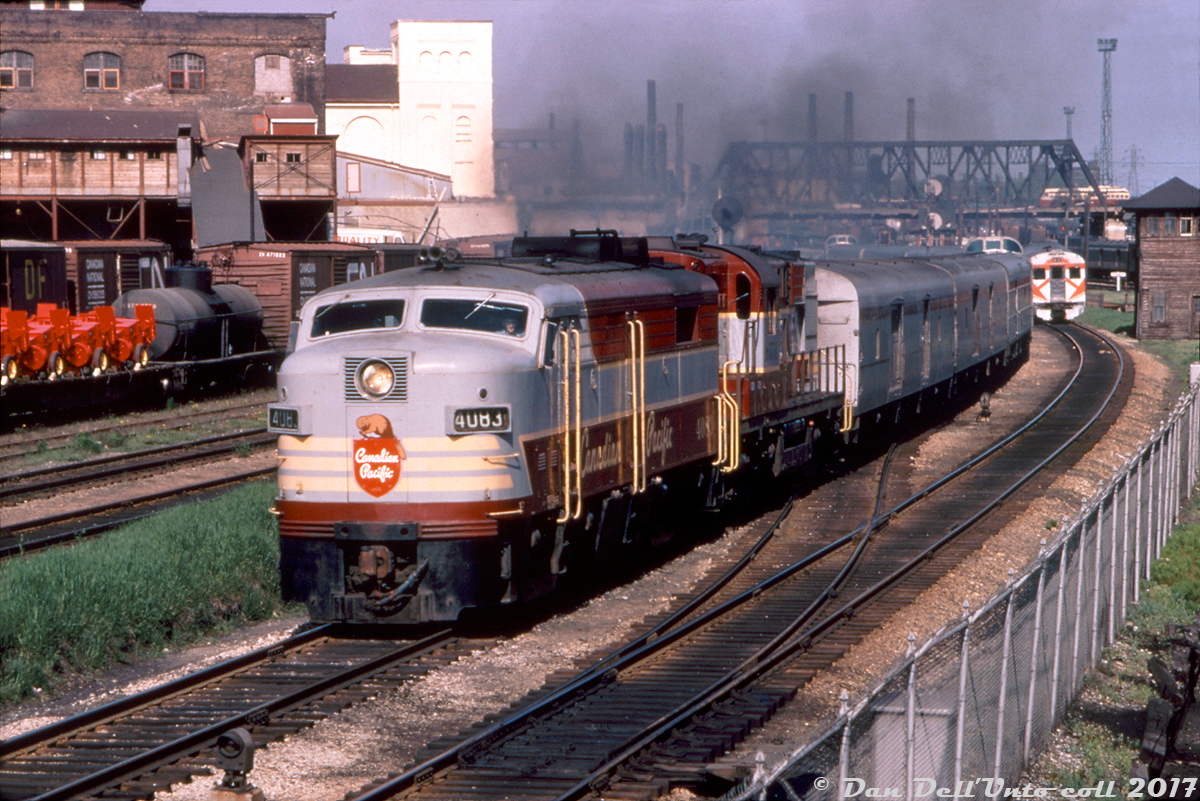 There's a lot of classic action in this picture perfect shot: Canadian Pacific train #11, the Toronto-Sudbury leg of CP's "The Canadian", departs downtown Toronto northbound on the Galt Sub with FPA2 4083 and an 8570-series RS10 heading up the stainless steel consist (with two regular steel baggage cars on the head end). The timetable departure time for #11 from Union Station is noted as 5:30pm, with the next stop being CP's West Toronto Station further up the line, before taking the connecting track over the diamond and heading up CP's MacTier Sub out of the city. The "hockey mask" scheme RDC car by CP's Tecumseh Street Tower is an employee shuttle from Lambton Yard to John Street terminal, held by Tecumseh St. waiting for rail traffic to clear before being lined inbound. Note the TTR's Cabin D interlocking tower even further back (visible behind the signals). In the background, CN S13 switchers and passenger cars can be seen around Bathurst Street bridge, along with a red and cream TTC PCC streetcar crossing the bridge northbound.To the left, a mix of CN freight is visible including noodle logo 40' boxcars, and a flatcar loaded with brand new red farm equipment from the nearby Massey Ferguson manufacturing plants around Strachan Avenue. Behind them is the Quality Meat Packers slaughterhouse (once the Toronto Municipal Abbatoir), that operated into the late 2000's. Lots of smokestacks from industry and lighting towers for the sprawling rail yards downtown litter the skyline.This shot was taken from the dirt mound at the south end of Parkdale Yard, which was used to divert runaway cars into, in order to prevent them from rolling downgrade into the downtown area.John Freyseng photo, Dan Dell'Unto collection (duplicate slide).