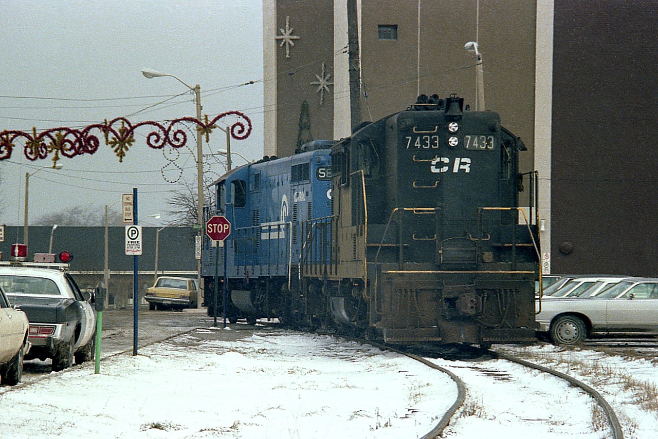 Back in the days when it was the Conrail Mainline thru Niagara Falls, we see two CR units having just entered the CR-CN connector which went over to the CN yard in town. This is a 'going away' shot, with CR 5823 leading 7433, about to cross thru the intersection of Queen and Erie Sts.  That is Roseburgs Dept store on the right, still in business and sporting holiday decorations. In the far background left and centre is the Bridge St. VIA station. The power will head over to the CN, and then bring some traffic back to CR Montrose at the south end of the city. Quite the old time lights on the cop car.