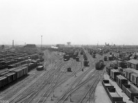 An overview of Canadian National's sprawling Mimico Yard is pictured looking westward from the east end yard tower back in 1954. There is a large abundance of classic steam-era equipment visible, including tons of both wooden and steel 40' boxcars, old wooden vans, an Alco S-series diesel switcher working one of the leads, and wisps of white smoke from steam engines working in the yard. The RIP track and car shops are visible on the right, and the leads to the roundhouse are present on the left (the roundhouse and its smokestack are visible on the left in the distance, with the ice house for servicing ice-cooled refrigerator cars nearby). The city water tower in New Toronto (still standing today) is visible off in the distance by Kipling Avenue, as is the old National Silicates plant in the distance on the right. The Oakville Sub mainline tracks originally ran past Mimico on the northernmost edge of the yard (hidden on the far right beyond the yard tracks). <br><br>Mimico Yard, formerly CN's main west-end freight yard in the Toronto area at the time, has undergone many significant changes over the years. The transition from steam to diesel in the 50's made a lot of the old steam-era facilities redundant, and the Mimico roundhouse was demolished in parts between 1965-1969 (the old coal tower managed to survive into the 80's however). The most notable was when CN opened its new Toronto Yard (later renamed MacMillan Yard) north of the city in Concord/Maple, shifting much of its freight yard operations there and vacating a good chunk of Mimico Yard on the north side to allow government-owned commuter agency GO Transit to establish its base of operations out of the old CN car shop buildings, forming the start of Willowbrook Yard (more modern maintenance facilities were constructed on-site and opened in 1980). Also changed around the same time, the Oakville Sub mainlines were re-aligned right through the middle of the yard (cutting right through the middle of this image), and the former mainlines removed except for a service track to switch customers along the north side of the yard. Much of the yard tracks on the left were also removed for a new freight shed on the south-east side built around 1968.<br><br>Another significant change was when VIA moved from its Spadina roundhouse & yard facilities in downtown Toronto to their new Toronto Maintenance Centre in the mid-80's, built in Mimico Yard on the south side of the mainline, on the site of the old ice house. Over time as local customers dwindled and passenger services expanded, GO Transit and VIA Rail have taken over more and more of Mimico Yard. Today CN only occupies a small portion along the south side by New Toronto Street, used by locals for servicing the remaining freight customers in the area.<br><br>A similar present-day view can be had looking west off the Islington Avenue overpass, that was built to the west of the car shops over the middle of the yard around the early 1980's.<br><br><i>More Mimico images:</i><br>The Mimico coal tower and CNR Northern 6167: <a href=http://www.railpictures.ca/?attachment_id=26895><b>http://www.railpictures.ca/?attachment_id=26895</b></a><br>Mimico Roundhouse ready tracks teaming with steam engines: <a href=http://www.railpictures.ca/?attachment_id=15340><b>http://www.railpictures.ca/?attachment_id=15340</b></a><br>CNR 6201 stored at Mimico awaiting scrapping: <a href=http://www.railpictures.ca/?attachment_id=29996><b>http://www.railpictures.ca/?attachment_id=29996</b></a><br>First-generation diesel power at the east end of Mimico: <a href=http://www.railpictures.ca/?attachment_id=18441><b>http://www.railpictures.ca/?attachment_id=18441</b></a>
