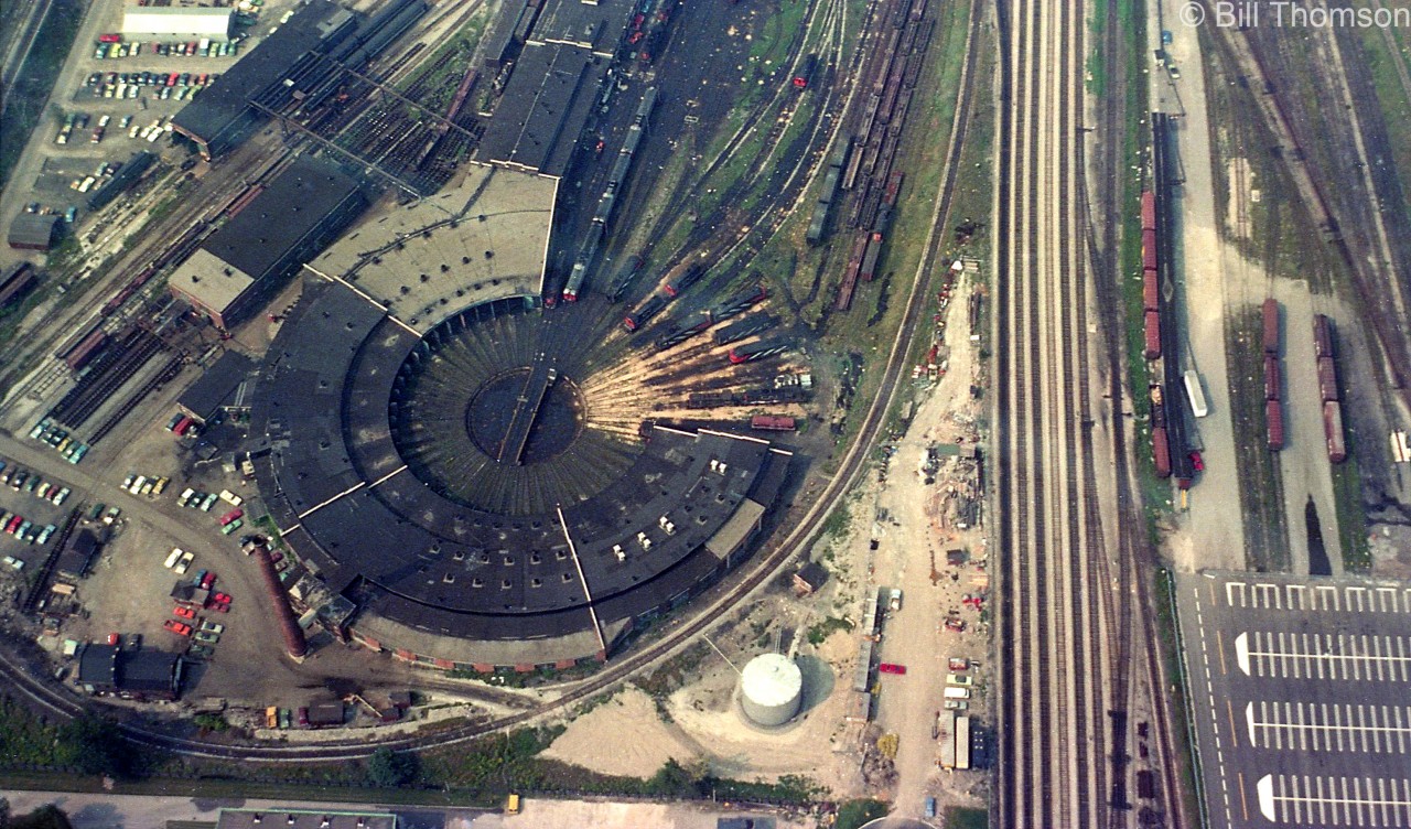 CN's Spadina Roundhouse is seen looking down from the CN Tower. The usual variety of power is visible including CN zebra-striped FP9's and FPA4's, S13 switchers for the coachyard, RDC cars and even a GP40 by the roundhouse. Steam generator cars and piggyback flats also populate the tracks nearby. The tracks to the right is the Toronto Terminals Railway corridor leading to Union Station, and to the right of that are boxcars spotted at CN's downtown team tracks and loading platforms. The CN Tower parking lot below was built over part of this area. The "Loop Track" where passenger equipment and consists were turned is visible around the bottom of the roundhouse.

In the upper left corner are CN's Stores buildings (note all the wheels), coach repair and paint shop facilities, as well as the "High Line" (bypass tracks) just in the corner (the Gardiner Expressway is further out of frame). Spadina Avenue overpass (where a lot of railfan photos of Spadina have been taken from over the years) is just out of the picture across the top.

Today, this view would be of the Skydome (Rogers Centre) that was built on the side of Spadina Roundhouse in the late 80's.