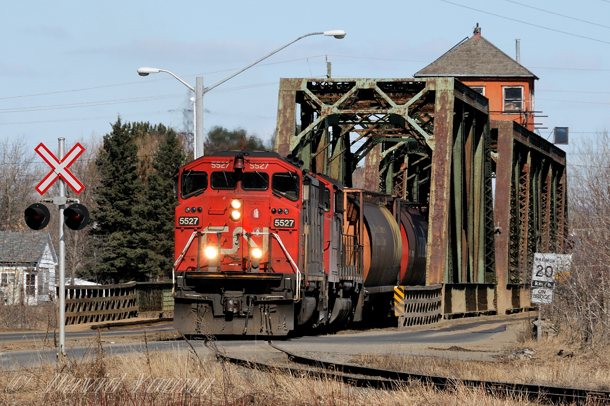 Rolling across the old swing bridge over the Kaministiquia River, utilizing power off the nights road freight from Fort Frances of SD60F 5527 and GP40-2wl 9525, the 07:30 Neebing Yard crew has grain loads for Mission Terminals Ltd and Cargill on the drawbar. There duties will be pull and spot the elevators and may have had a switch at the Bowater Sawmill, all located at the end of the spur.This bridge is a rail/ road bridge, built by the Grand Trunk Pacific Railway in 1906. A contract was signed between the GTP and the Corporation of the Town of Fort William and a sum of $50,000 dollars was given to the railway. In return the municipality gained "the perpetual right to cross said bridge for street railway, vehicle and foot traffic on roadways supported by brackets on each side of the railway bridge." On October 29, 2013 the bridge burned, repairs were made to continue rail traffic but the bridge has since remained close to vehicle and pedestrian traffic due to a dispute between CN and the City of Thunder Bay determining who is responsible to pay for repairs. The issue had been taken to court and in June 2017 a judge ruled CN does not have to fix the bridge. The dispute continues as in August 2017 Thunder Bay appealed the decision. This is the most direct link for people commuting between the City and the Fort William First Nation.