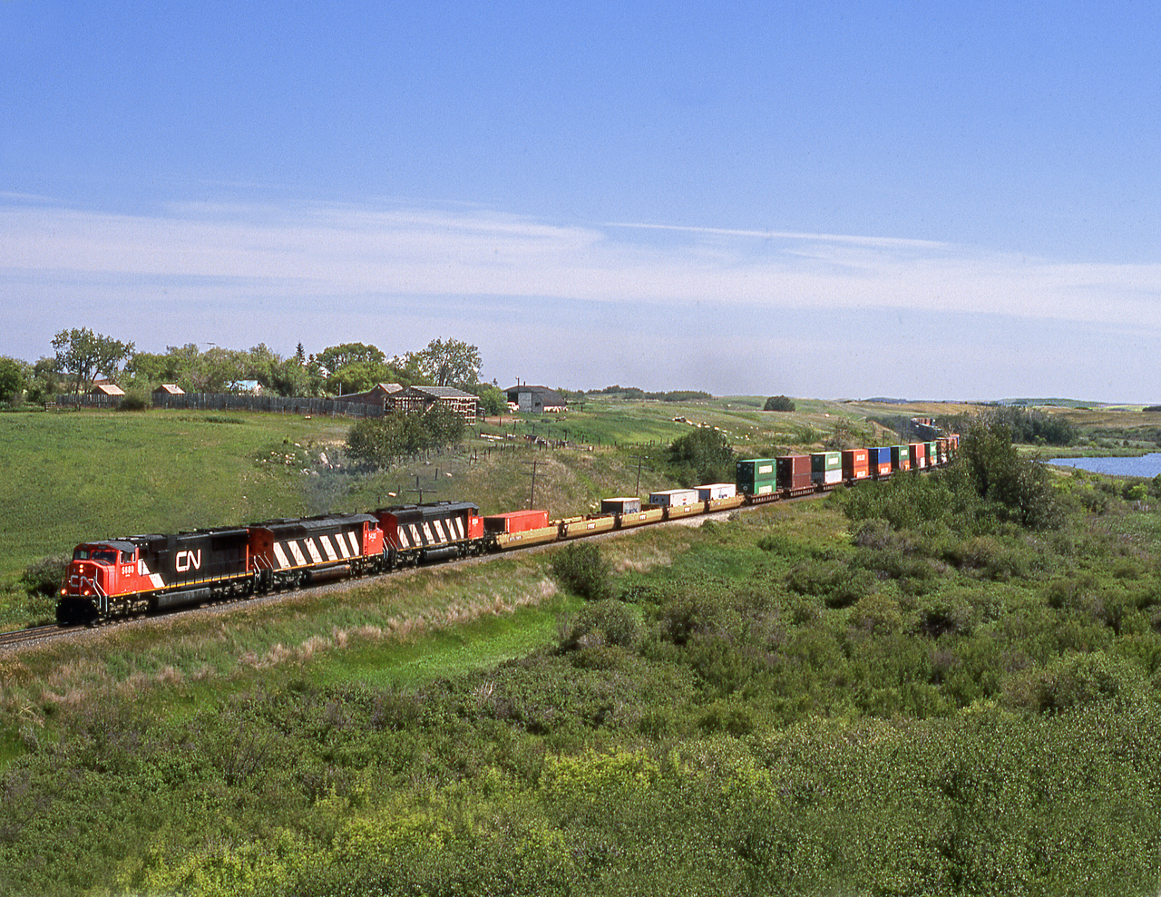 Westbound train 169 rolls through a coulee between Neola siding and crew change point of Biggar. CP's parallel Wilkie Sub. is on the top of the valley behind the farmyard