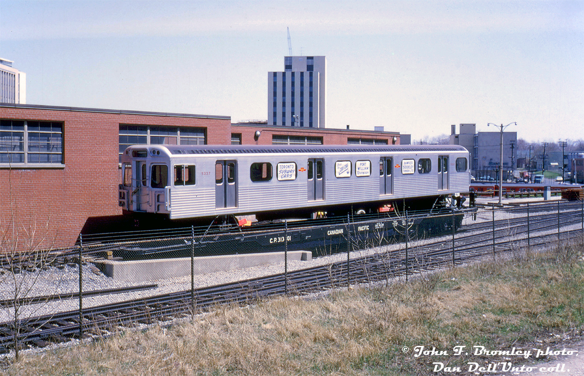 Brand new Toronto Transit Commission H1 subway car 5337 is shown on Canadian Pacific flatcar 313001, delivered by CN inside of the TTC's Davisville Yard at the unloading ramp waiting its turn to roll onto the TTC's subway tracks for the very first time.   TTC ordered 164 new subway cars from Hawker Siddleley Canada for use on the then under-construction Bloor-Danforth subway line in 1964. They would be built at the old Can-Car plant in Thunder Bay, Ontario, and numbered after M1's 5300-5335 built a few years earlier by Montreal Locomotive Works (for the University line).  TTC H1's 5336-5499 were the first of a long line of subway cars built at the Thunder Bay plant (now owned by Bombardier), and 5336-5337 were the first "married pair" to be delivered: 5336 delivered the previous day (May 4th), and 5337 delivered May 5th as shown. They were also the only two H1's delivered to Davisville Yard, as the rest of the H-series cars were delivered to the new Greenwood Yard via a rail spur off CN's Kingston Sub. The H1's went on to see a good three decades in service on the Yonge-University-Spadina and Bloor-Danforth lines until the mid-late 90's, when the worn-out fleet were retired by new T1 subway cars built at the same plant the H1's were built at three decades earlier. While most were sold for scrap, a few made their way into work car service including 5336 and 5337.  The flatcars used to deliver these cars were interesting themselves: CP 313000-313007 were built from old chopped-down heavyweight sleeping cars, and equipped with deck rails and side support pedestals for loading and transporting subway cars. According to photos I've been able to find, three were initially done in the early 60's for delivery of the MLW M1's (418103, 418124, 418125), and by the H1 deliveries were joined by the eight 313's in 1965, until removal from the roster in 1976. Later H4 and H5 deliveries in the 70's were done with North American Car NIFX 1000-series flatcars.  A number of "standard gauge" tracks and sidings ran in the south-west part of Davisville Yard here, entering from a switch off of CN's Belt Line Branch (ex-Grand Trunk, originally built as the Toronto Belt Line Railway). New subway cars (including the Gloucesters, MLW M1's, the first two H1's, and even work car RT-10 "Tokyo Rose") and supplies were delivered to Davisville from the 1950's when the yard was built, until around 1969 when the Belt Line Branch east of the new Spadina Expressway was abandoned (its rails were pulled up beginning in April of 1970). The old right-of-way was sold to the City of Toronto a few years later and converted to a walking trail, which it remains today (the Kay Gardiner Beltline Trail).  John F. Bromley photo, Kodachrome from the Dan Dell'Unto collection.