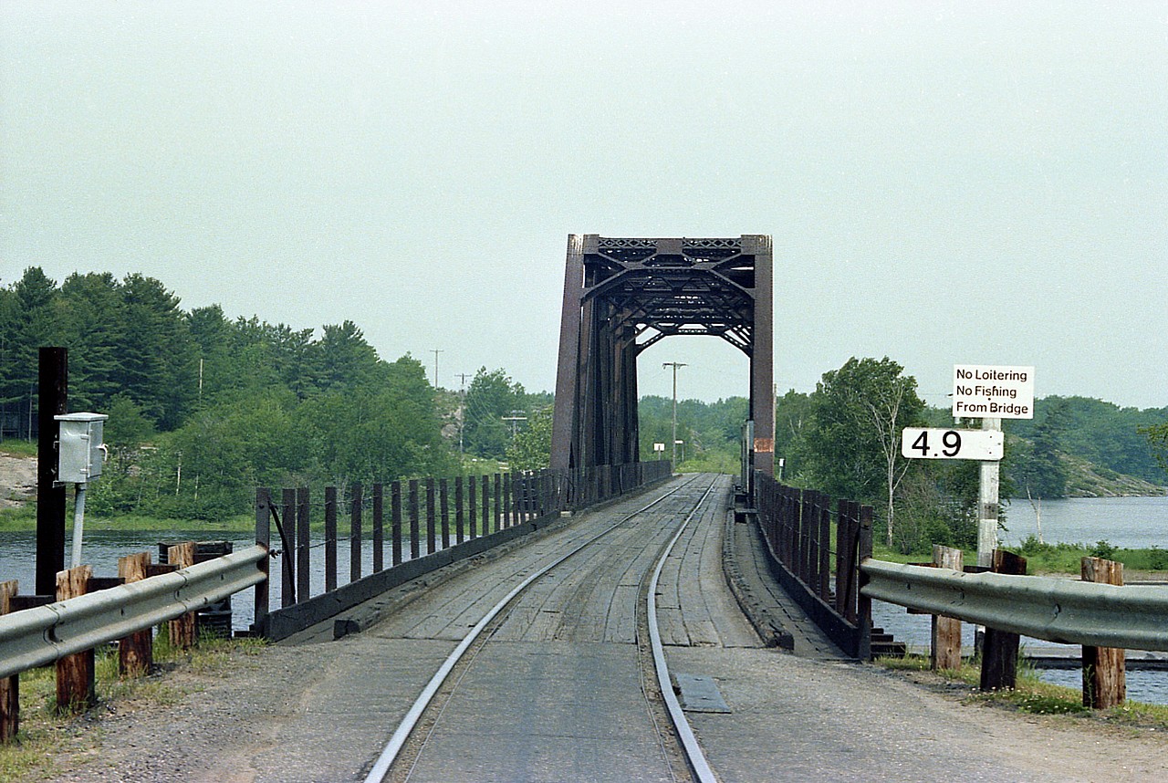 Depot Harbour was founded by 'visionary' John R. Booth back in 1892. The railroad connection was the Ottawa, Arnprior & Parry Sound Railroad (OA&PS). Became part of the Grand Trunk in 1904 and later, part of the CNR network. Shown is the road/rail bridge to Depot Harbour, which was situated on Parry Island, home of the Wasauksing First Nation. The town had a fascinating history, but it is all gone now; the last resident having left in 1980. At its best, the community had around 1600 residents, as well as a couple of grain elevators,warehouses, an excellent deep water pier and a well laid out residential area. The railroad was very busy, lake ships from Chicago and Milwaukee brought lumber, wheat and coal, which were unloaded onto the railcars and sent to Ottawa and Montreal and points East. A train every half hour. Eventually it all died out as better shipping routes were found. In 1926 the railroad, streamlining somewhat, closed the roundhouse, turntable, freight shed and offices and centralized activity at nearby South Parry. Very little happening any more and the town dwindled down to about 200 residents. On the night of August 14, 1945, mysteriously a blaze started in one of the grain elevators, used for the storage of cordite which was involved in the making of dynamite at the nearby Nobel Canadian Industries Ltd (CIL). The building exploded and took most of the town with it in a raging inferno. Jobs gone, people left. The opening of the St. Lawrence Seaway in 1959 assured Depot Harbour it would never amount to anything ever again.
All of this is involved in why this pictured Wasauksing Swing Bridge existed. As far as I know it was the only railroad / auto combined bridge in Canada. Trains had to stop and crew checked for approaching vehicular traffic on both sides before advancing over the bridge. And the bridge often was opened for boat traffic. Quite the all-purpose structure. And only one lane !!!! This image is from nearly 40 years ago, and 10 years after this photo was taken, the last train left Depot Harbour and the tracks you see here were removed. A neat piece of Canadian railroad history.