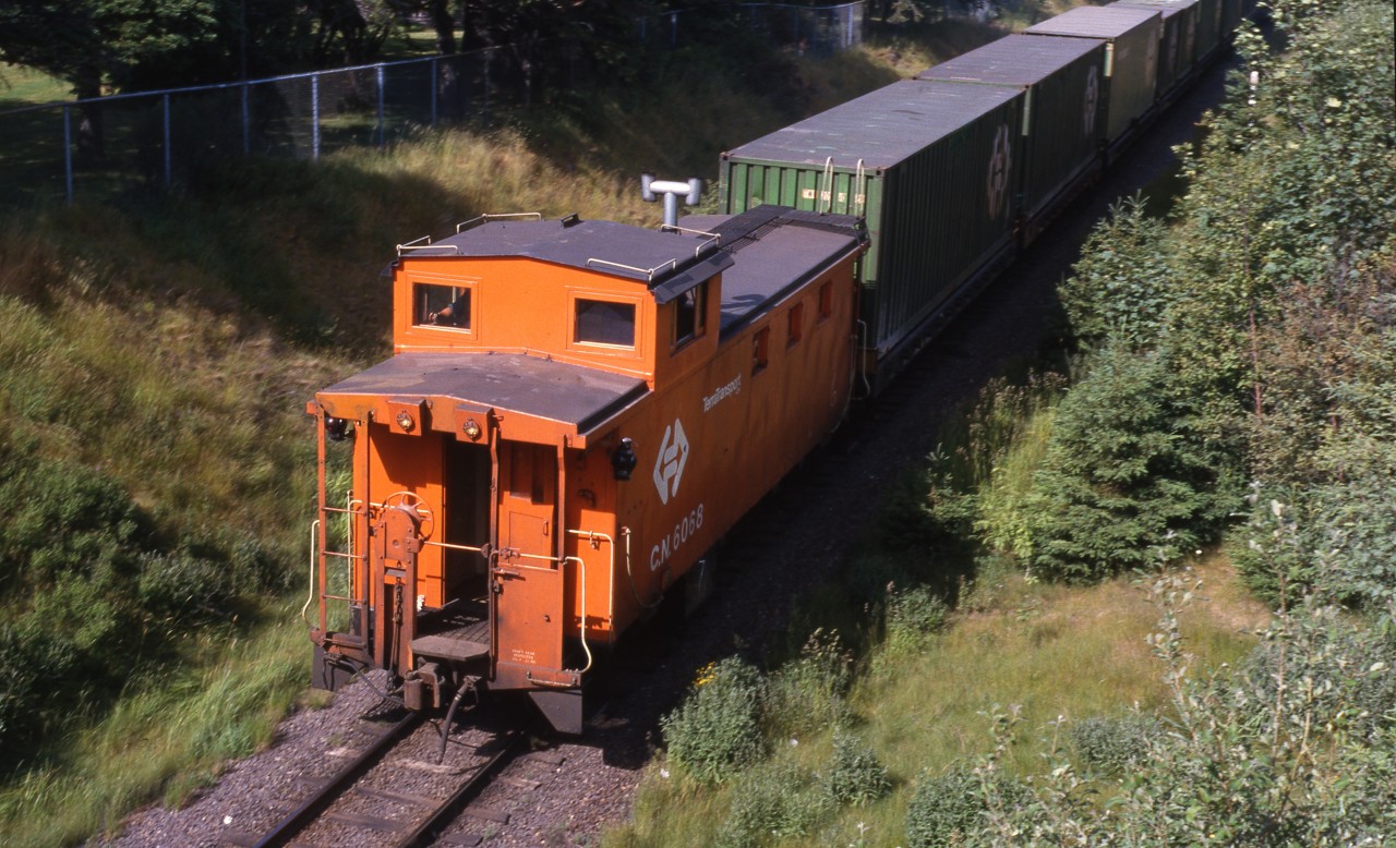 TERRA TRANSPORT TAIL END. Narrow gauge TerraTransport Caboose 6068 brings up the rear of Extra 926 East (see image 13831) at the Bowring Park pedestrian overpass on the beautiful Sunday afternoon of July 19, 1987. This all-steel caboose was one of five built in 1967 by National Steel Car of Hamilton, Ontario and had all welded construction. It was from a total order of twenty built from 1961 to 1967 in five lots by that company to replace a variety of old wooden cars. Fortunately it survived the shutdown a year later and wound up as a playhouse at the McDonalds Restaurant in Bay Roberts before being moved to the grounds of the Avalon North Wolverines Search and Rescue Unit. In September of 2017 it was bought by rail buff Ken Mercer and moved to his property at Bristol's Hope, Conception Bay and restored to its former glory where it sits today.