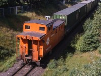 TERRA TRANSPORT TAIL END. Narrow gauge TerraTransport Caboose 6068 brings up the rear of Extra 926 East (see image 13831) at the Bowring Park pedestrian overpass on the beautiful Sunday afternoon of July 19, 1987. This all-steel caboose was one of five built in 1967 by National Steel Car of Hamilton, Ontario and had all welded construction. It was from a total order of twenty built from 1961 to 1967 in five lots by that company to replace a variety of old wooden cars. Fortunately it survived the shutdown a year later and wound up as a playhouse at the McDonalds Restaurant in Bay Roberts before being moved to the grounds of the Avalon North Wolverines Search and Rescue Unit. In September of 2017 it was bought by rail buff Ken Mercer and moved to his property at Bristol's Hope, Conception Bay and restored to its former glory where it sits today.