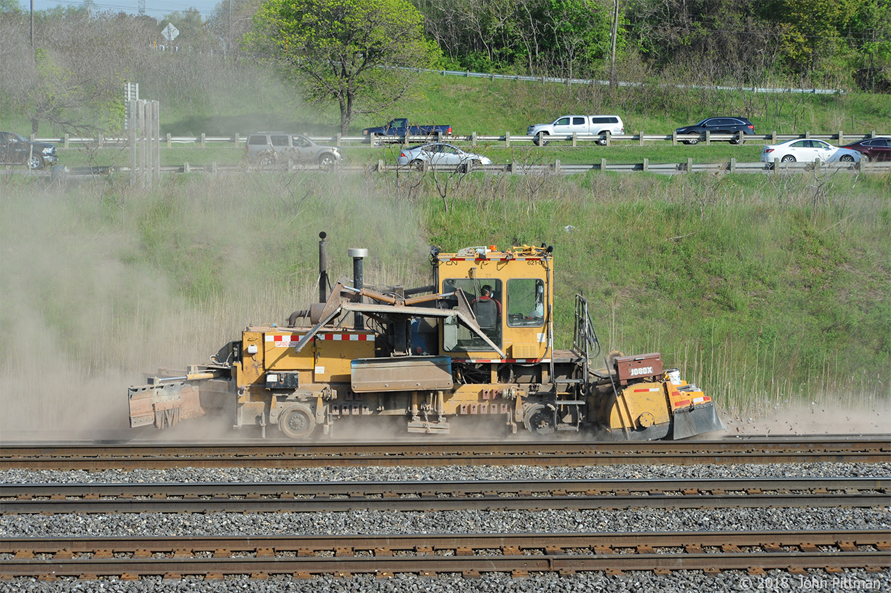 Multi purpose MOW machine CN 621-01 in a cloud of dust and spitting stones is on a mission to keep the ballast below the tie-tops on the recently re-ballasted Aldershot West yard lead. Note the operator wears a dust mask.  
After my next visit to this location, I found 2 dog ticks had climbed up onto my head - watch out in long grass. Restrain ticks with clear adhesive tape and identify -> http://www.tickencounter.org/tick_identification/tickid_nonflash