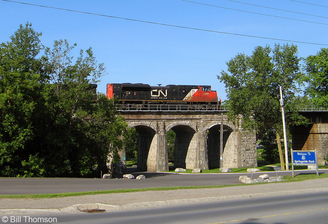 CN SD70M-2 8891 pops out between the trees heading eastbound on the Kingston Sub over the stone-arched Napanee Viaduct, built in 1856 to cross the Napanee River.