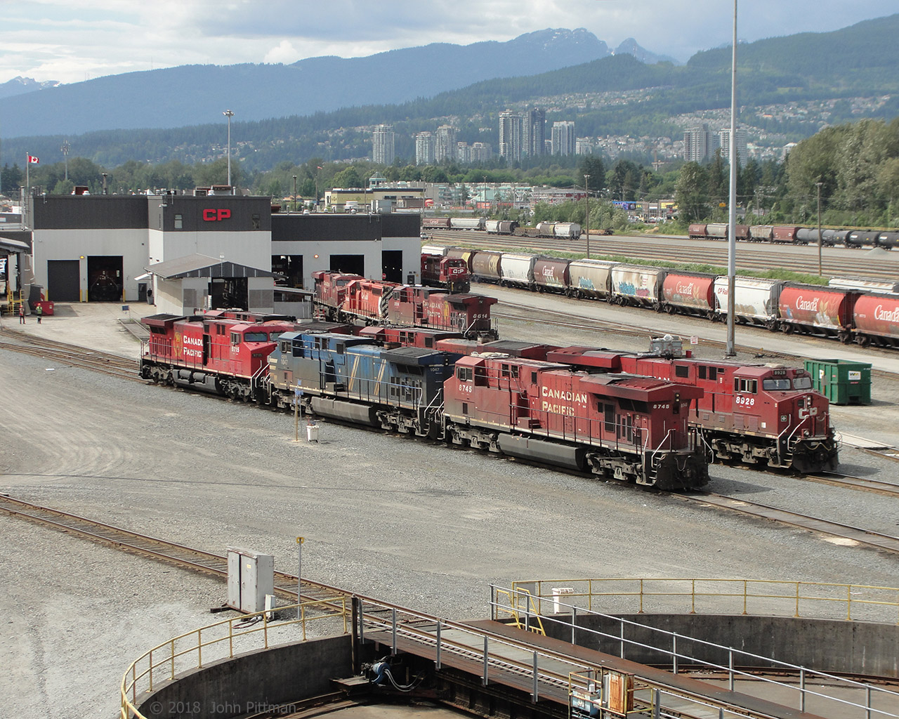 A north-west facing view of CP's Port Coquitlam yard diesel shop and turntable.  In the background part of the city of Port Coquitlam, with some of Coquitlam further up the hillside, and coastal mountains beyond. Nice scenery for a railyard.  Vantage point is the sidewalk of the Coast Meridian Overpass (bridge). 
In the nearest row are AC4400's CP 9715 and CEFX 1047, then ES44ac CP 8745.  A pair of CP AC4400's and ES44ac CP 8928 are in the next row.  Closer to the diesel shop are AC4400 CP 8614, an SD40-2, and an ES44ac. Looks like GP38ac CP 3011 beside the shop.