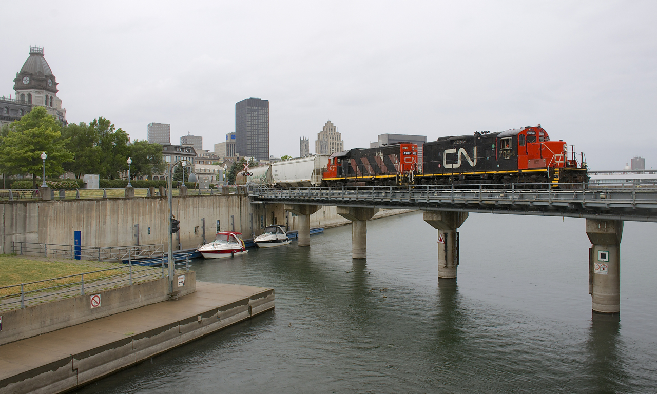 With some much needed rain falling, a transfer leaves the Port of Montreal with CN 7054 & CN 4707 leading a 12-car train over the Lachine Canal.
