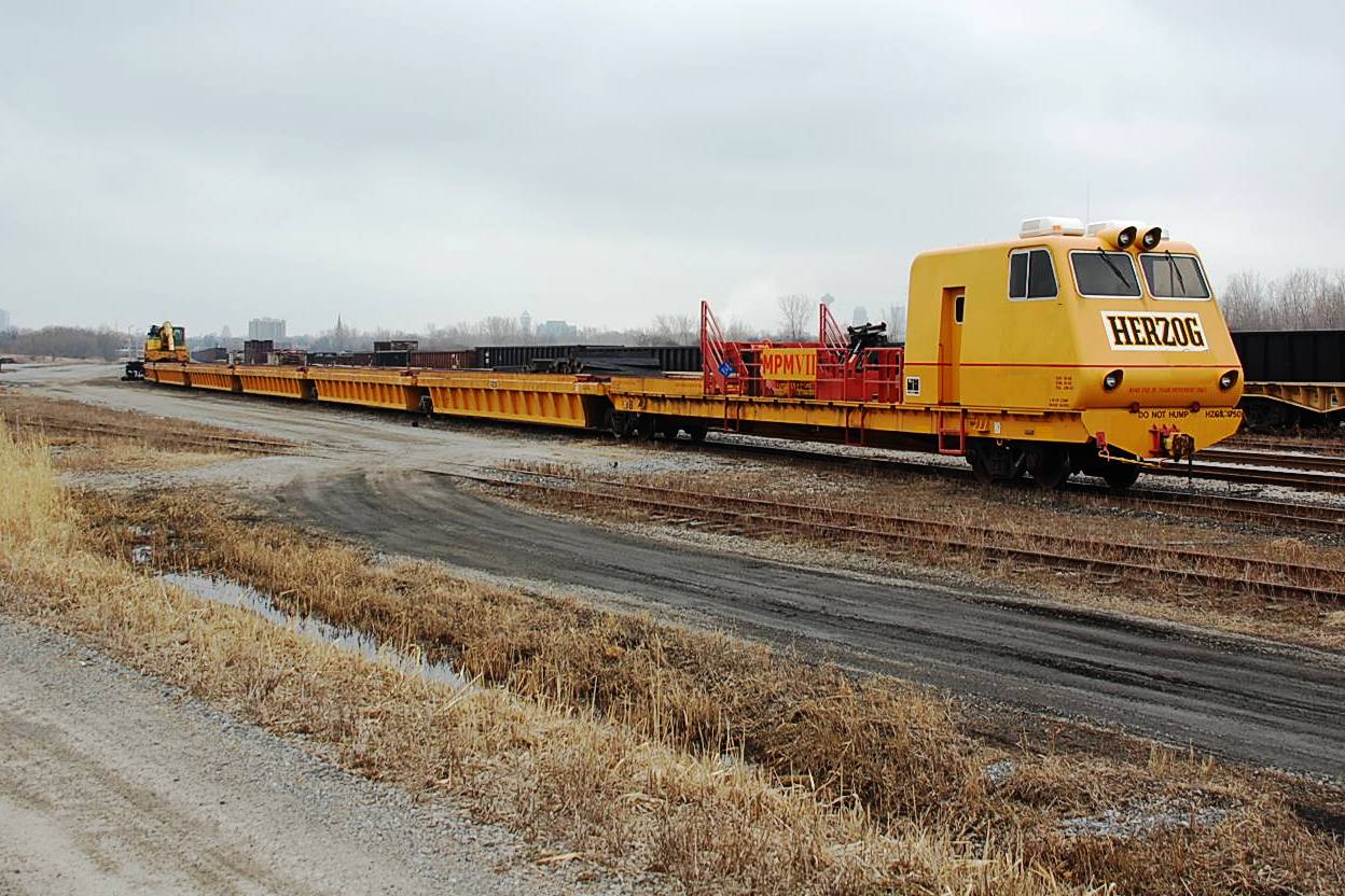 An otherwise overcast and dreary Sunday morning in March yielded a bit of excitement when I came across this elusive 'beast' sitting in the Niagara Falls yard. This multipurpose work unit (including a back hoe/loader that can travel along it's own gondola cars) is Herzog 175, MPM VII. Difficult to photograph due to it's unusual length, it was captured by my camera in three stages. 
Some landmarks visible in the background include the spire of the Saint Patricks church on Victoria Ave, Casino Niagara (ex-Maple Leaf Village) tower, and the Skylon tower.