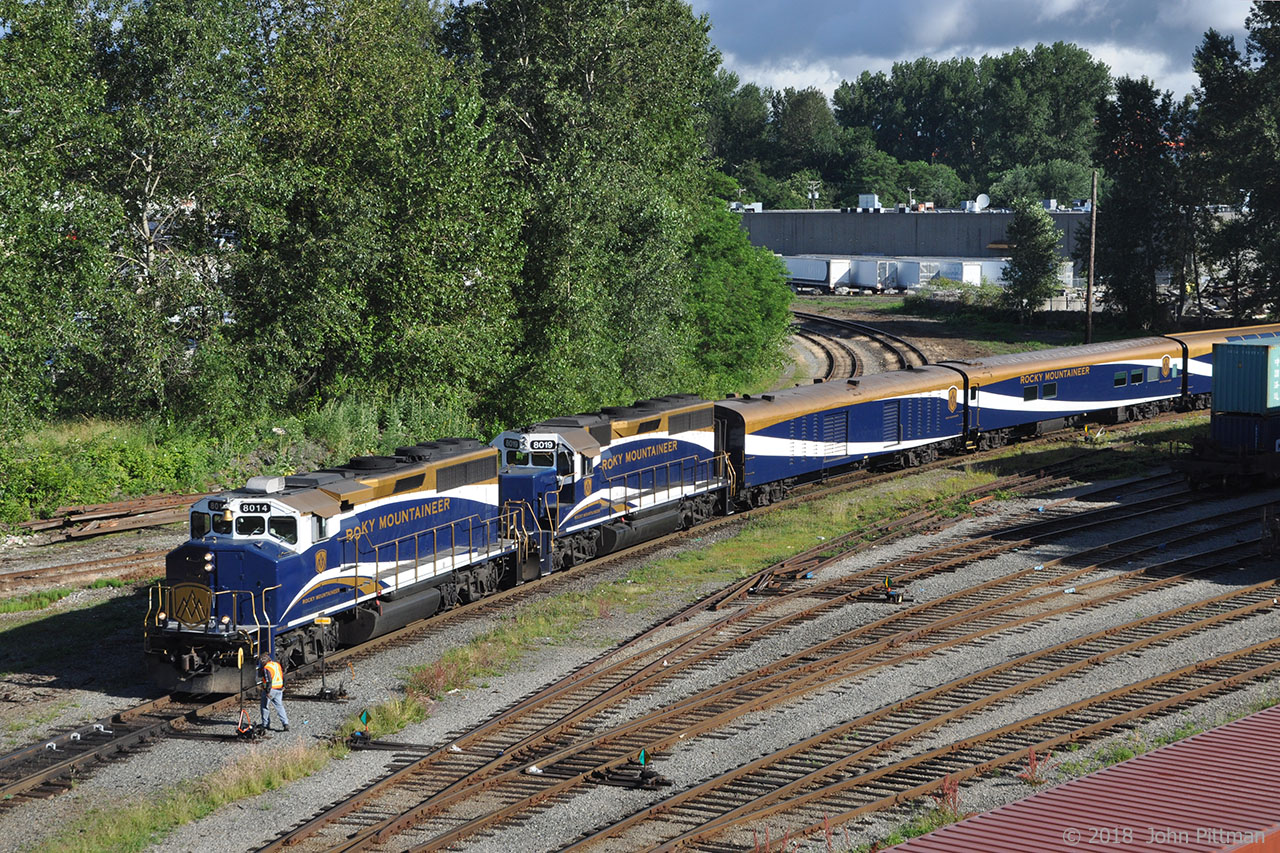 Halted at the end of a reverse move from Rocky Mountaineer Railtour's Vancouver station to wye their train, ex-CN GP40-2W RMRX 8014 leads the June 25th departure with rebuilt GP40-2 RMRX 8019 (origin Penn Central). This long train includes HEP coaches at both ends, and RMR's silver and gold class coach accommodations for both a CN and a CP route tour.  Similarly to VIA, CN, and CP, RMR follows CN/CP directional running in the Fraser Canyon as far as Kamloops.  At Kamloops RMR splits their eastbound trains into CN and CP sections. Westbound RMR trains arriving from CN and CP routes are merged at Kamloops for the trip to Vancouver. 
Switches at Burrard Inlet Junction (a.k.a. the Diamond) have been set so that the train can proceed forward on the CN/BNSF New Westminster subdivision. 
The Rocky Mountaineer Railtours station is on the north side of CN's downtown rail yard.  It was built within the shell of a former locomotive maintenance building that once serviced CN Passenger trains. Before RMR's current station site was renovated for their needs, their trains used Pacific Central station.