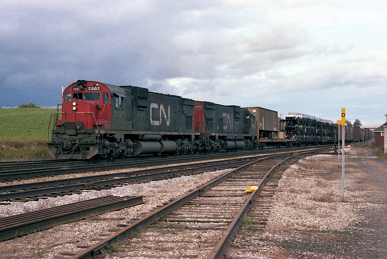 I'm appreciating the "D" derail post in this image. For otherwise, even the guys who hang out at Copetown by the mill might not catch on immediately that this is where the photo was taken many years ago. CN 2007 and 2009 a pair of MLW C-630Ms head westbound in this cloudy day shot past what is almost a barren landscape compared to what is there today.  Note the long gone MoW shack on the right, and the Hwy 52 overpass in the extreme background.