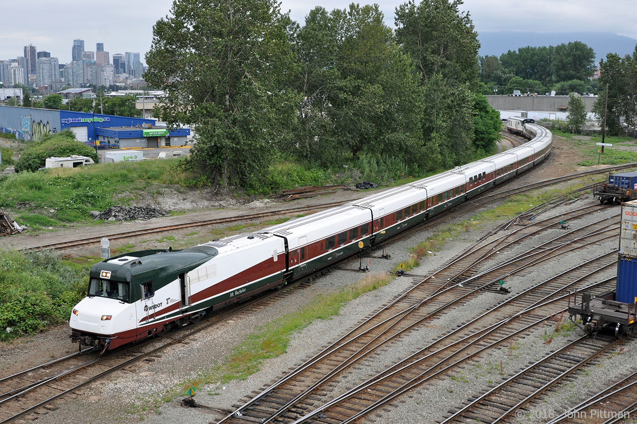 Amtrak Cascades train 519 is seen departing from track leading to Vancouver's Pacific Central Station, with Talgo Pendular Series 8 coachset "Mt Bachelor" and its integrated Control Cab/HEP leading - temporarily.    Scheduled to depart at 5:45 PM, it was passing here at 6:30 PM within a mile of the platform. 
Most fans consider that the integrated Talgo Control Cab/HEP is NOT one of the better-looking objects intended to lead a train.  I've heard it compared to the face of Donald Duck, or a snail without its shell.  Talgo coach sections are designed low-slung for cornering performance.  A higher cab floor level was required by the 2 axle lead truck, plus having the cab higher than the roof-line of the coaches improves visibility.  (Only place I've seen more than 1 axle in a Talgo coachset).  The unit number is hard to read, low-contrast black digits near the back of the brown swoosh. There are no illuminated number boards, which is rare. 
On this occasion the delayed departure was because of insurmountable technical difficulties with driving the train from the Control Cab. The solution was to wye the train using the CN / Rocky Mountaineer downtown yard.  Siemens SC-44 "Charger" locomotive AMTK 1403 will be in the lead when they enter the BNSF/CN New Westminster sub at CN Junction after freight movements had cleared.  The control cab has both headlights and red tail lights illuminated, which seems abnormal.